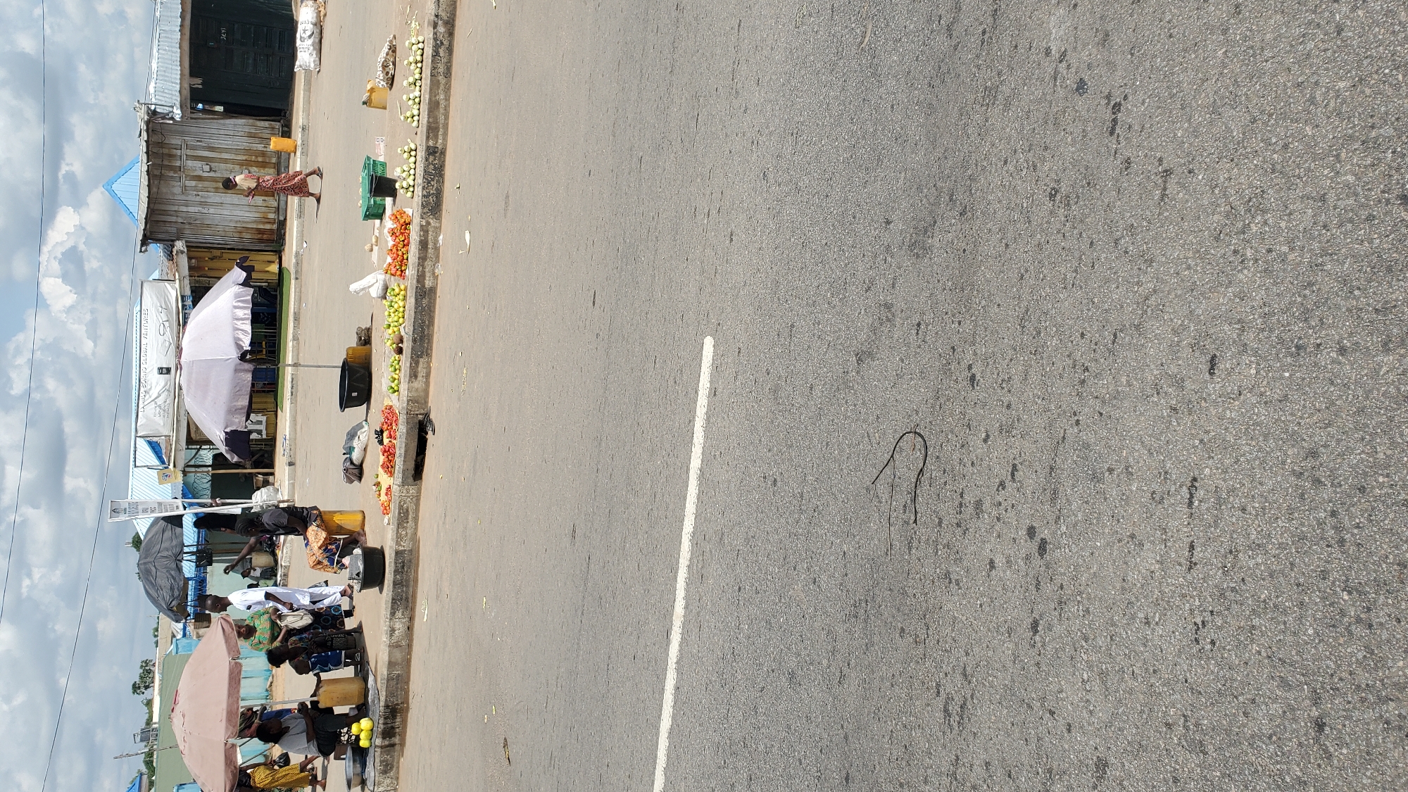 In Jalingo, Nigeria, a street scene unfolds under a partly cloudy sky, showcasing a bustling market. Several individuals are engaged in commerce, with women seated and tending to their wares, which include a variety of fresh produce like tomatoes and onions. Umbrellas provide shade for the vendors and their goods. The market is situated alongside a paved road, marked with a white line, suggesting it's a roadside setup. In the foreground, a dark, coiled object lies on the asphalt, its identity unclear from the image. The background reveals a row of small shops and stalls, indicating a commercial area.