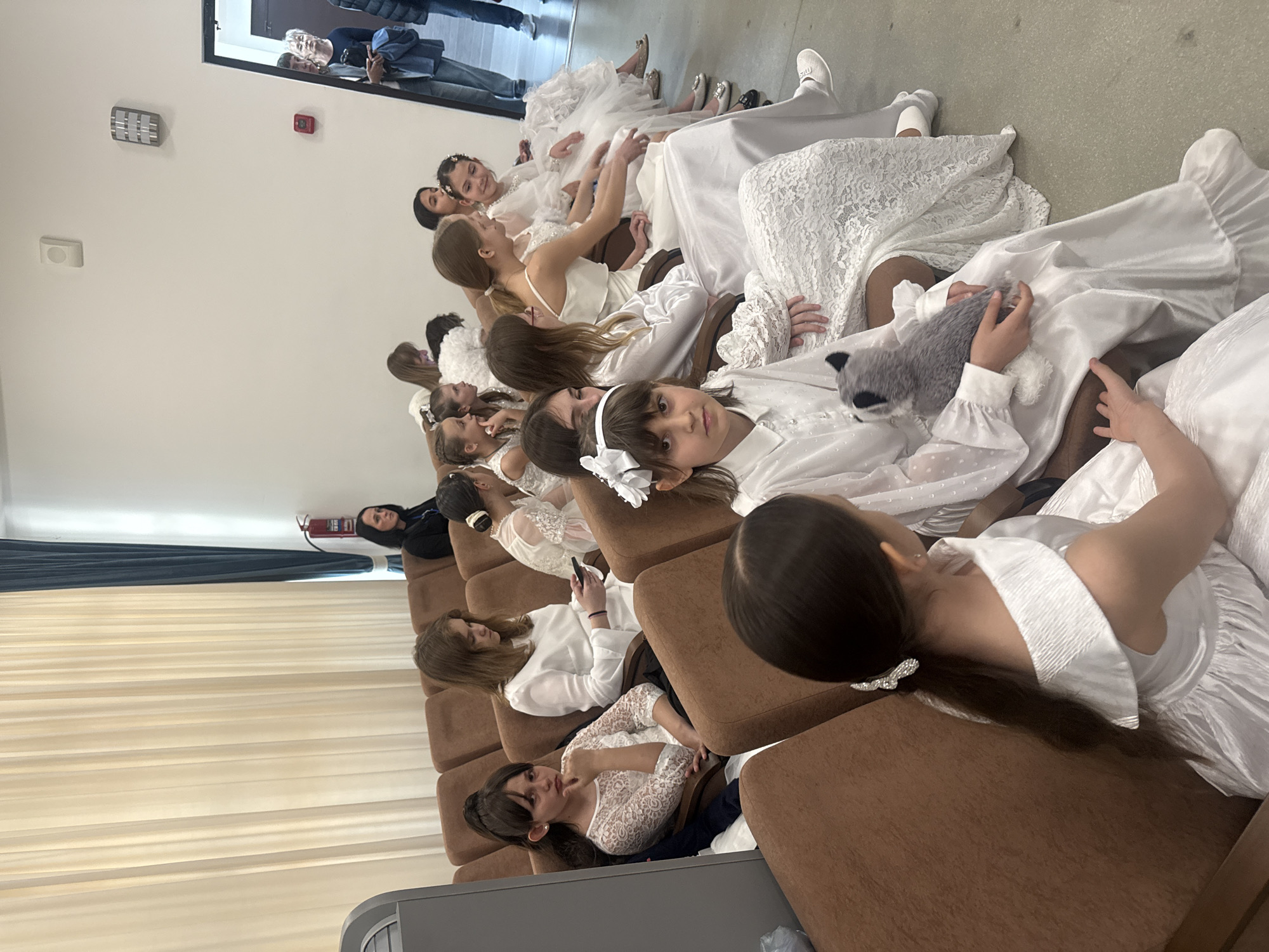A medium, eye-level shot depicts a group of young girls, possibly attending a special event, seated in a row on brown cushioned chairs against a white wall. The event occurs indoors. They are all dressed in white dresses, varying in style and fabric. Some girls have white bows in their hair. One girl is holding a grey plush animal in her lap. In the background, there is a curtained window, and a woman in black clothing is visible. At the far end of the room, there is a mirror reflecting individuals who seem to be observing the scene. A fire extinguisher is mounted on the wall near the window. The location appears to be Unknown, Unknown.