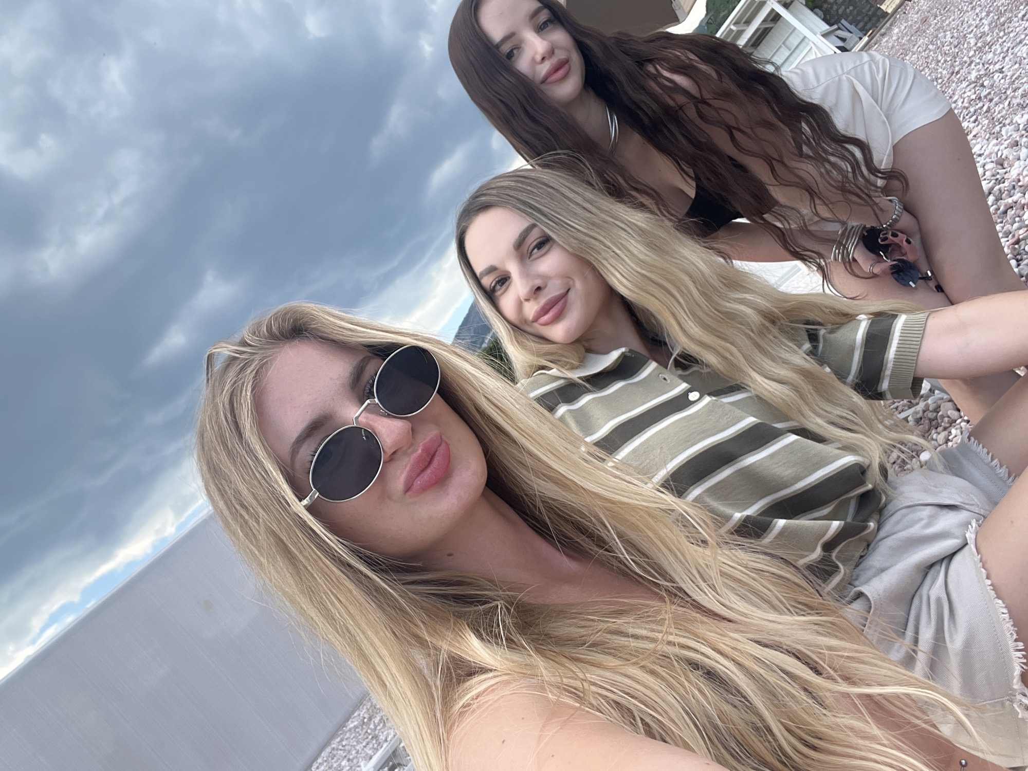 A selfie image shows three women posing outdoors in Sveti Stefan, Montenegro. The foreground woman has long blonde hair, round sunglasses, and full lips. The woman in the middle has blonde hair, and wears a striped olive green and white collared shirt. The woman in the background has long, dark brown hair and wears a black top and white shorts. They are on a pebbled beach with white structures in the background, partially obscured by cloudy sky. The image is taken at an upward angle.