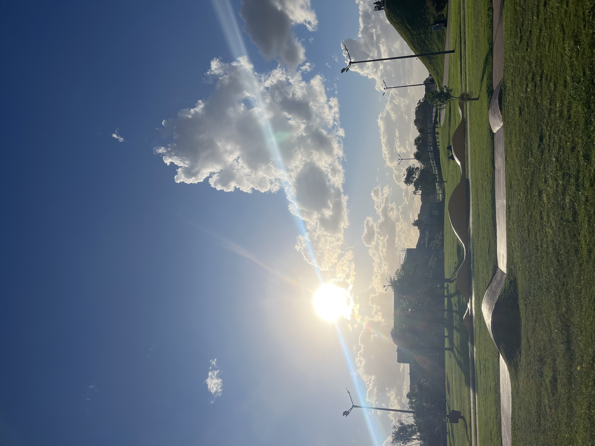 The image shows a bright, sunny day at Parque de la Memoria in Buenos Aires, Argentina. The sky is predominantly blue with scattered cumulus clouds, with the sun shining intensely, creating lens flare. The park features grassy areas interspersed with curving concrete pathways and wave-shaped sculptures. Streetlights are visible in the background, suggesting the park's urban location. The light is strong, creating sharp shadows on the grass and pathways. No people are visible in the image.
