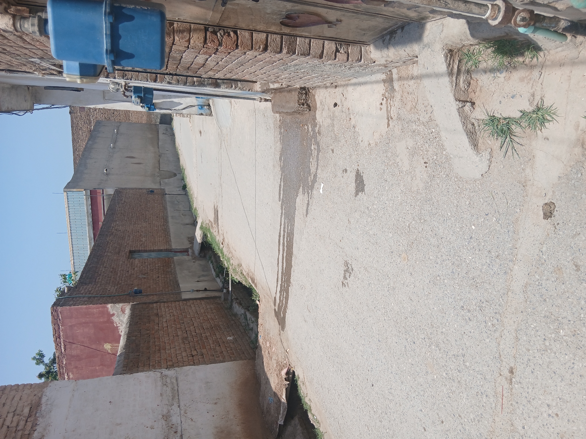 The image shows a narrow, paved street in Peshawar City Tehsil, Pakistan, bordered by brick and concrete buildings. The street appears to be a residential or commercial alleyway. A blue gas meter is prominently displayed on the exterior of a brick building on the right side of the frame, with pipes and electrical conduits also visible. Other buildings exhibit a mix of brickwork and plastered surfaces. A small ditch runs along the side of the street, with some vegetation growing in it. The sky is clear and blue, indicating a sunny day. The scene is devoid of any people or visible activity.