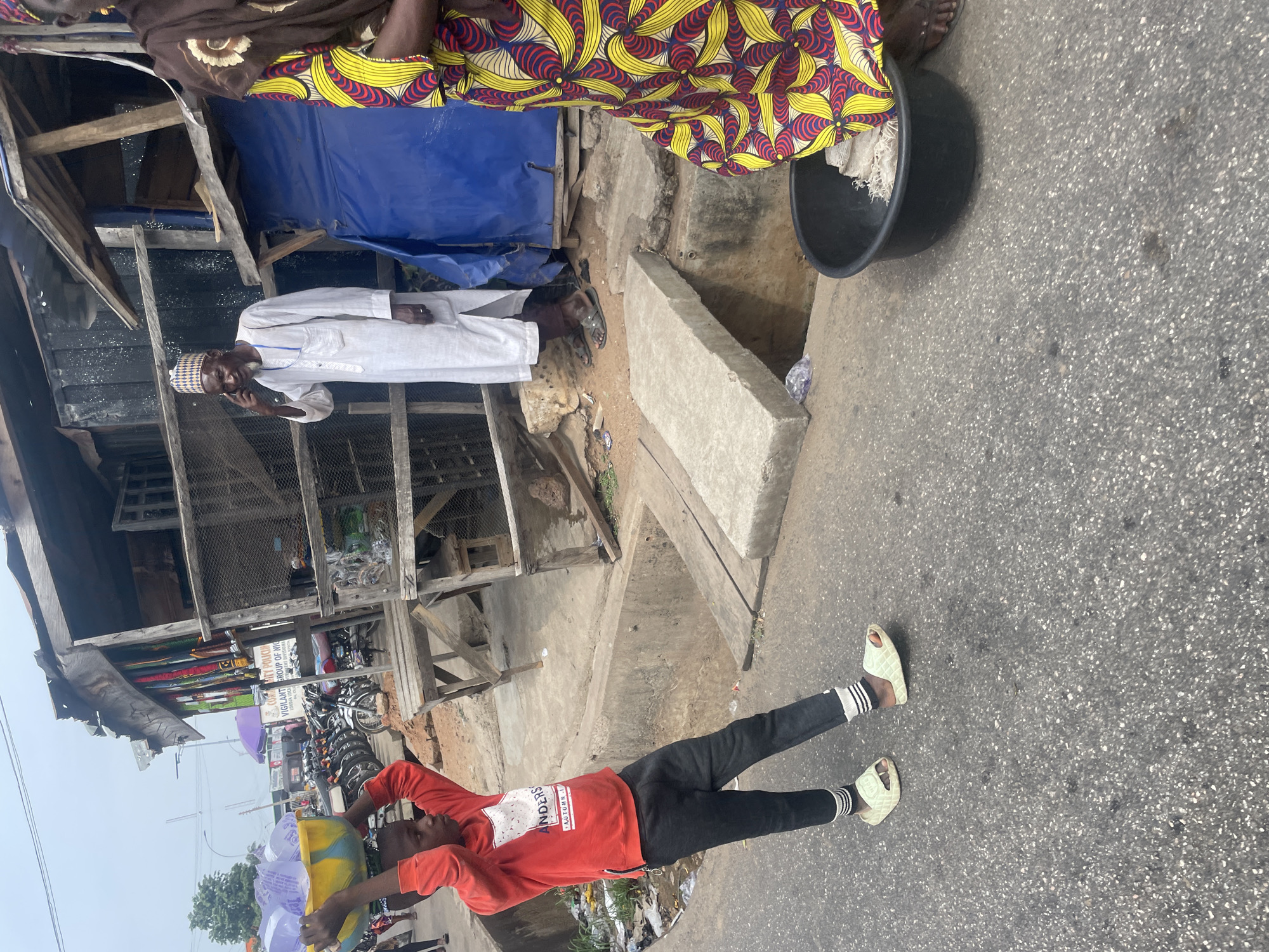 The image shows a street scene in Lokoja, Nigeria. A person in a red sweater and black pants is walking down the side of the road, carrying bags filled with yellow and blue items. An older man in a white robe and cap stands near a stall constructed of wood and corrugated metal. Another person wearing a colorful patterned garment and a woman’s brown garment stand near a black basin. Motorcycles are parked in the background near a sign that says “Key Polikoff Vigilante Group of Nigeria”. The surroundings appear to be cluttered with debris and the road is paved.