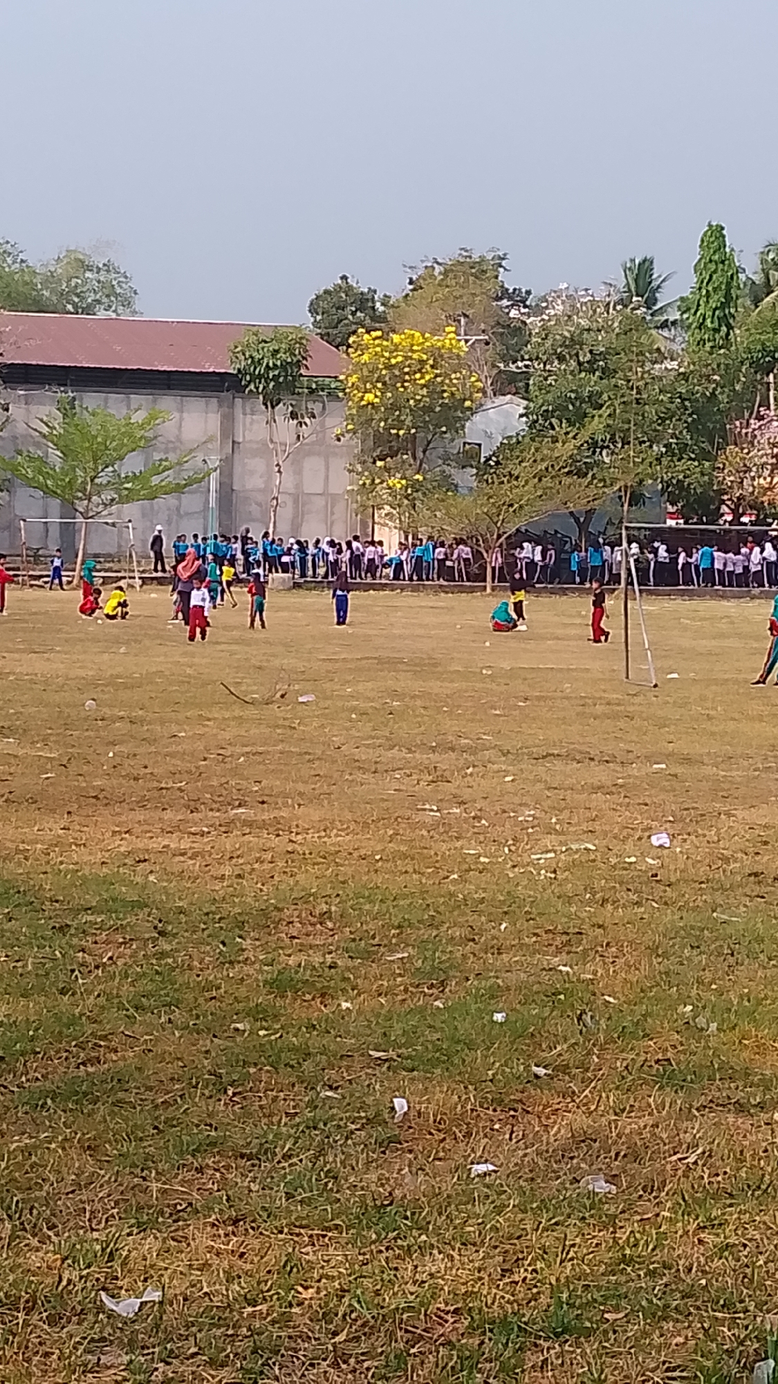 The image depicts an outdoor scene on a grassy field, likely a schoolyard or park, with a crowd of people in the background, many of whom appear to be students. The field itself is sparsely covered with dry grass and scattered with litter. In the mid-ground, several children are engaged in activities, with some appearing to play a game resembling cricket or a similar bat-and-ball sport. One child in the foreground is wearing a blue outfit, while another in the mid-ground is in a red and white outfit. A soccer goal is visible on the left side of the frame. Behind the field, a building with a reddish-brown roof is present, along with trees, including a prominent tree with yellow flowers. The sky is clear and bright, suggesting daytime. The context provided indicates this scene is located in Karanglangu, Indonesia.