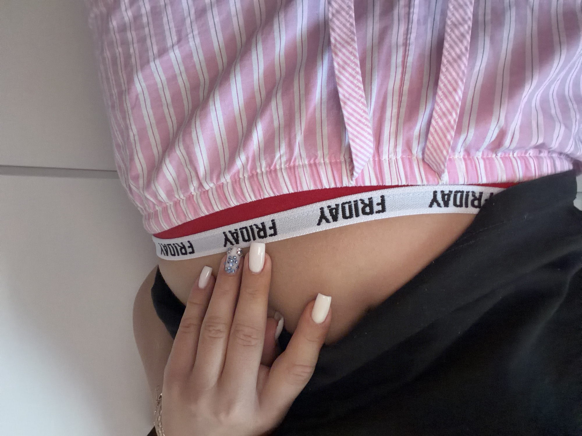 The image shows a close-up of a person's midsection, focusing on their attire. They are wearing pink and white striped pajama shorts with a white elastic waistband that has the word "FRIDAY" repeated on it in black lettering. A black garment, possibly a sweatshirt or sweater, is visible covering the upper part of their torso. The person's hand, with neatly manicured white nails and one nail decorated with rhinestones, is resting on their hip, partially obscuring the waistband. The background is a plain, light-colored wall. The image does not contain any recognizable landmarks or features that would allow for localization to Novi Iskar, Bulgaria, or any other specific geographical location.