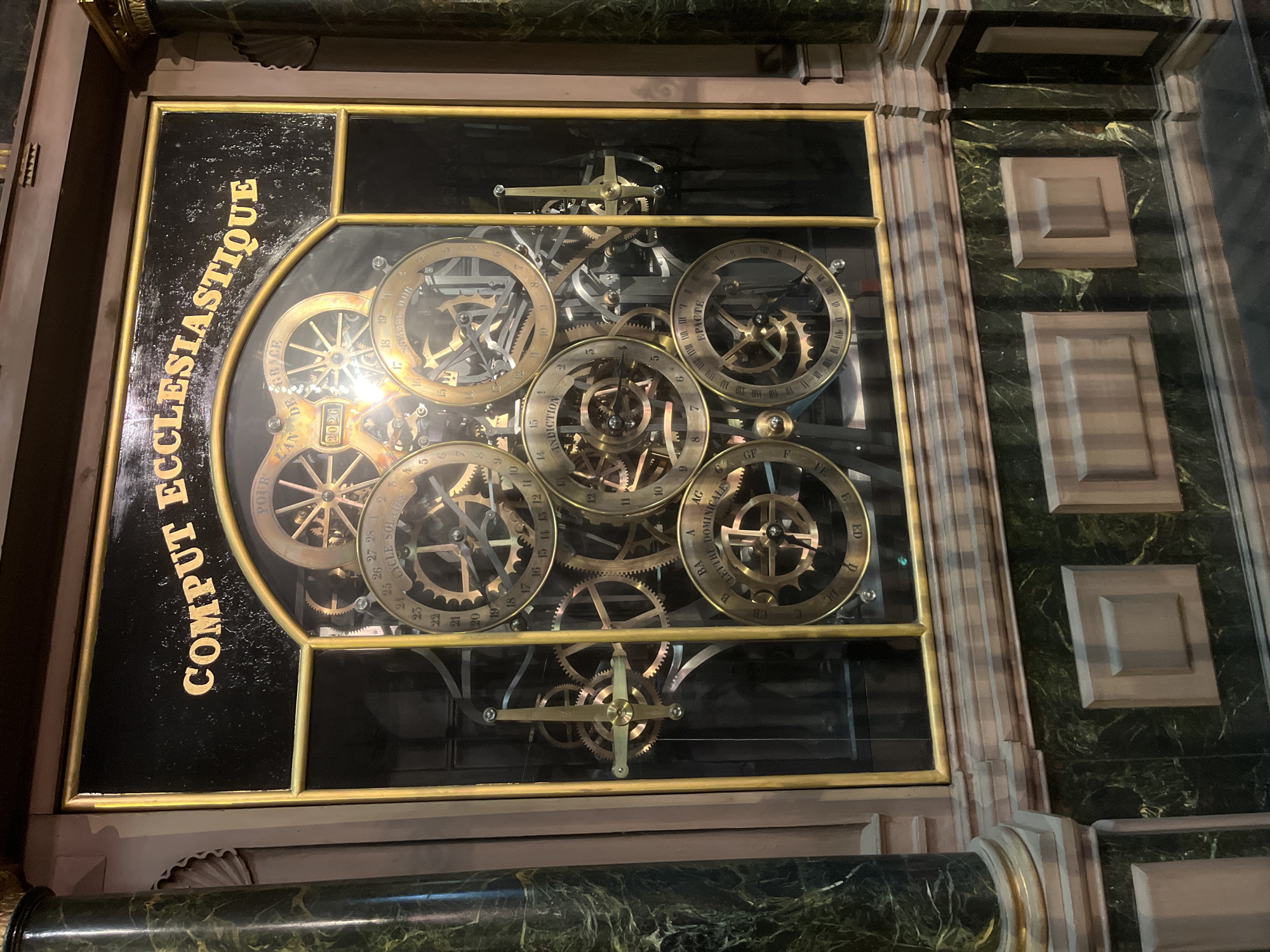 This media file showcases a close-up view of an intricate mechanical clockwork mechanism, likely part of a large astronomical or historical clock. The clock features multiple brass gears and rings, some with inscribed numbers and text. The text "COMPUT ECCLESIASTIQUE" is prominently displayed on a black background with gold lettering. Other visible text includes "POUR LAN DE" and "2026", indicating a calculation for the year. Several rings are labeled with terms such as "CYCLE SOLATRE", "INDICTION", "LETTRE DOMINICALE", and "EPACTE", suggesting the clock tracks various calendrical cycles. The gears are arranged in a complex, layered fashion, with some gears having visible teeth and others displaying Roman numerals or smaller subdivisions. The overall aesthetic is one of precision engineering and historical significance. The lighting appears to be artificial, possibly from an indoor setting, highlighting the metallic sheen of the clockwork. The surrounding structure of the clock is visible, featuring dark, possibly marble, panels with a green, marbled pattern and decorative moldings in muted tones. There are no people or clear indications of time of day or weather. The scene is static, focusing entirely on the detailed operation of the clock mechanism.