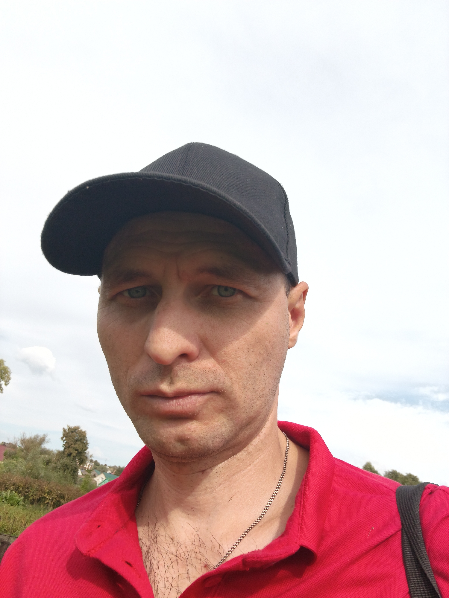 A close-up, outdoor, waist-up selfie shows a middle-aged Caucasian man wearing a black baseball cap and a red polo shirt. The man has fair skin, light blue eyes, and appears to have a slightly stubbly chin. His expression is neutral. He is wearing a silver chain necklace. Behind him, the background consists of a bright, slightly overcast sky and some greenery, including trees and what appear to be houses in the distance. The location is identified as Mokshan, Russia, suggesting an outdoor, possibly rural or suburban setting within that city.