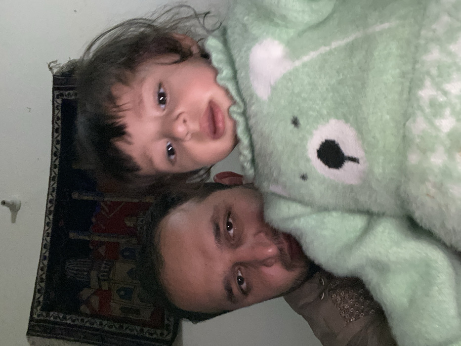 A rotated image shows a man and a young child. The man's face is near the bottom, with dark hair and a light brown complexion. The child, positioned above the man, has dark hair and is wearing a light green fuzzy bear-patterned outfit. A small rug with dark borders and red and brown designs is visible in the background. The background also features a plain white wall with a small fixture. The overall lighting is soft and diffused.