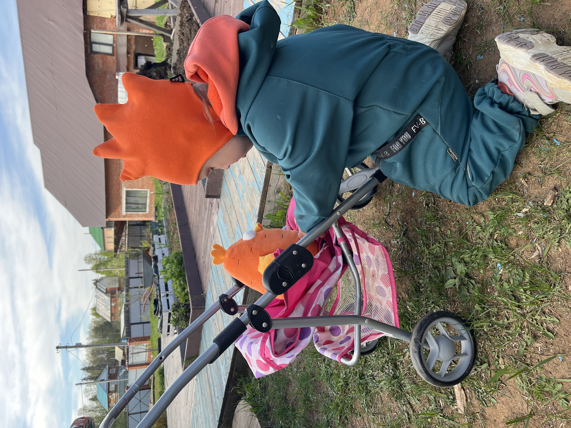 A child wearing a green jumpsuit with a bright orange hoodie and an orange hat is kneeling on the grass. The child is looking into a grey toy stroller that contains a stuffed, orange fish. The child is in the yard of a house in the city of Chaikovsky, Russia. Other houses are visible in the background. The grass is green and dry in some places.
