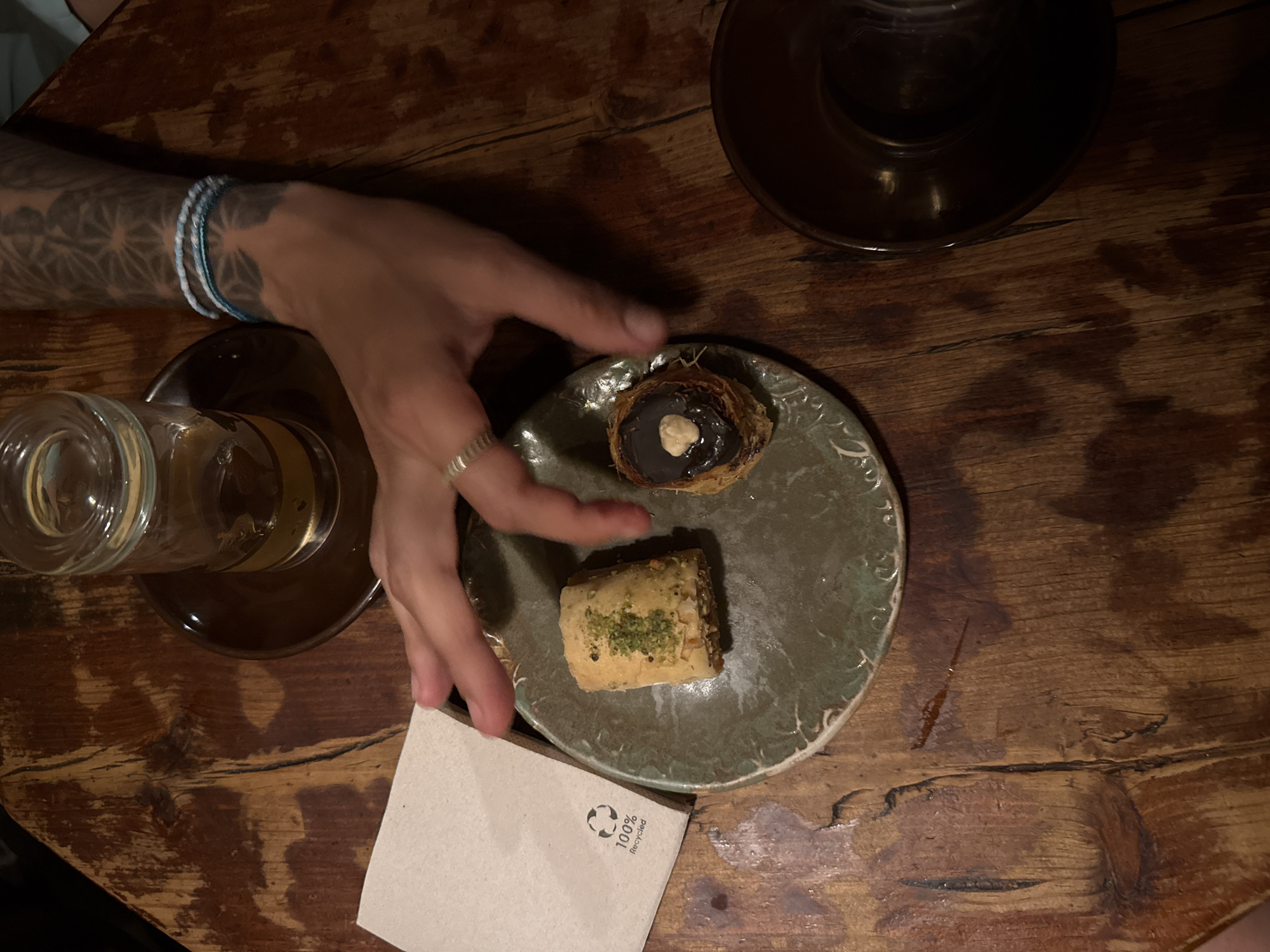 The image depicts a close-up, overhead view of a person's hand reaching for a small piece of baklava on a textured, green ceramic plate. The baklava appears to be topped with chopped pistachios. Next to it on the plate is another pastry, possibly a chocolate-covered bird's nest pastry with a nut on top. To the left of the plate is a folded white napkin with a "100% Recycled" symbol. In the background, on a rustic wooden table, is a clear glass with a dark liquid, likely tea, resting on a dark saucer. The person's hand, adorned with a silver ring and beaded bracelets, has visible tattoos on their forearm. The scene is set in Barcelona, Spain.