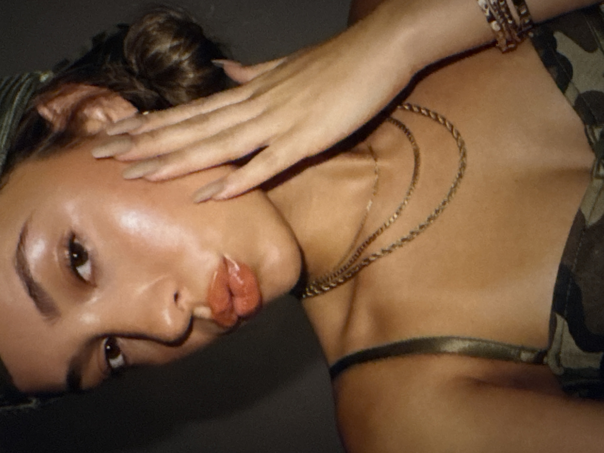 A close-up, overhead shot shows a woman's face with her hand on her cheek. She is wearing a green camouflage-patterned top and two gold necklaces. Her hair is pulled back into a bun. Her lips are glossy and appear to be puckered. The background is dark and out of focus. The image is lit with a warm light that highlights her skin. The location is not specified in Kaunas, Lithuania, but the image is an artistic portrait.