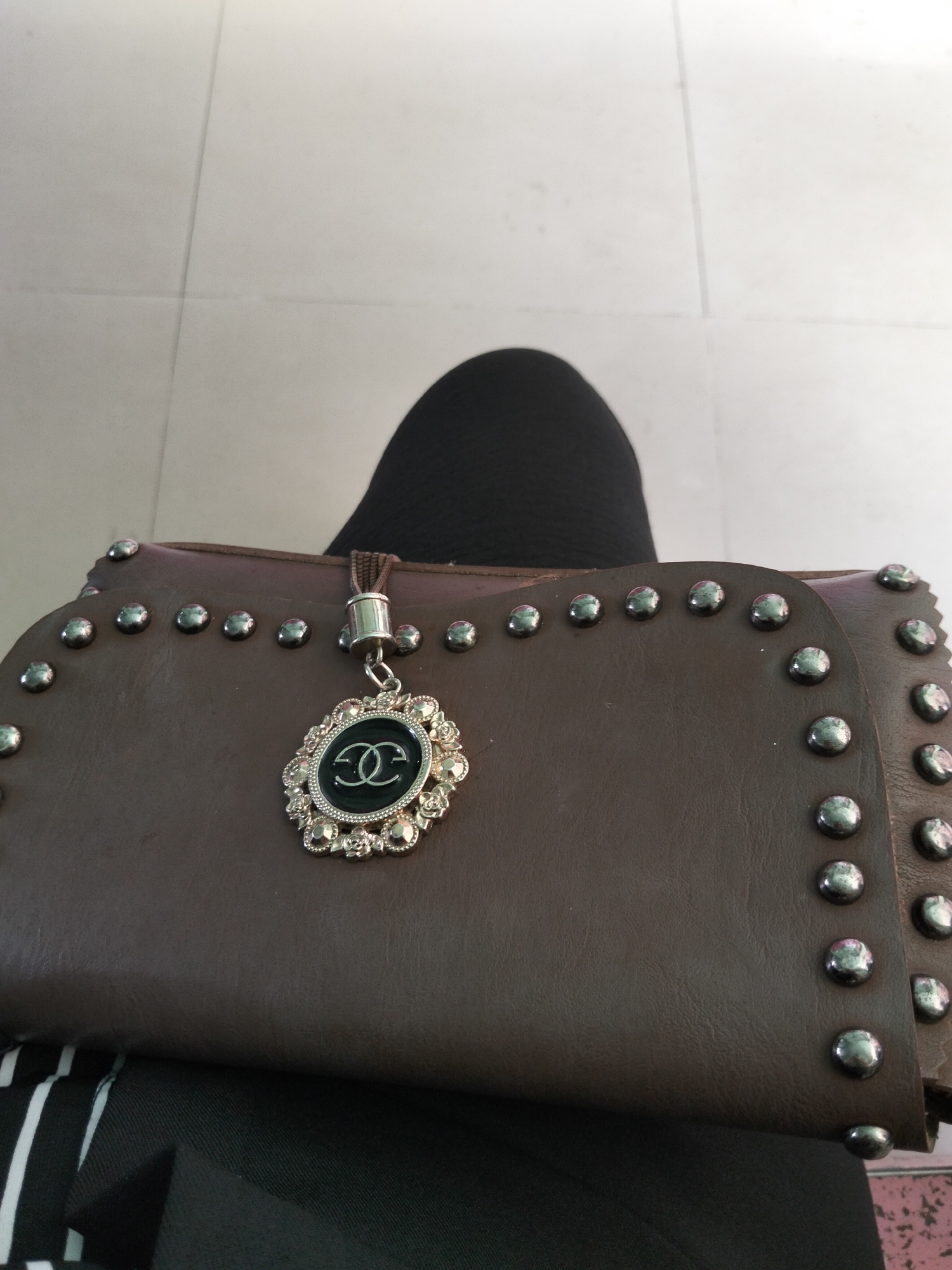 The image displays a close-up of a brown leather handbag adorned with numerous silver studs along its edges. A decorative charm, featuring a black oval with a double-C logo in silver, surrounded by a floral-patterned silver frame with embedded rhinestones, hangs from a small brown cord attached to the bag. Behind the handbag, a black fabric, likely part of clothing, is visible. The background consists of light-colored floor tiles with visible grout lines. The image appears to have been taken indoors.
