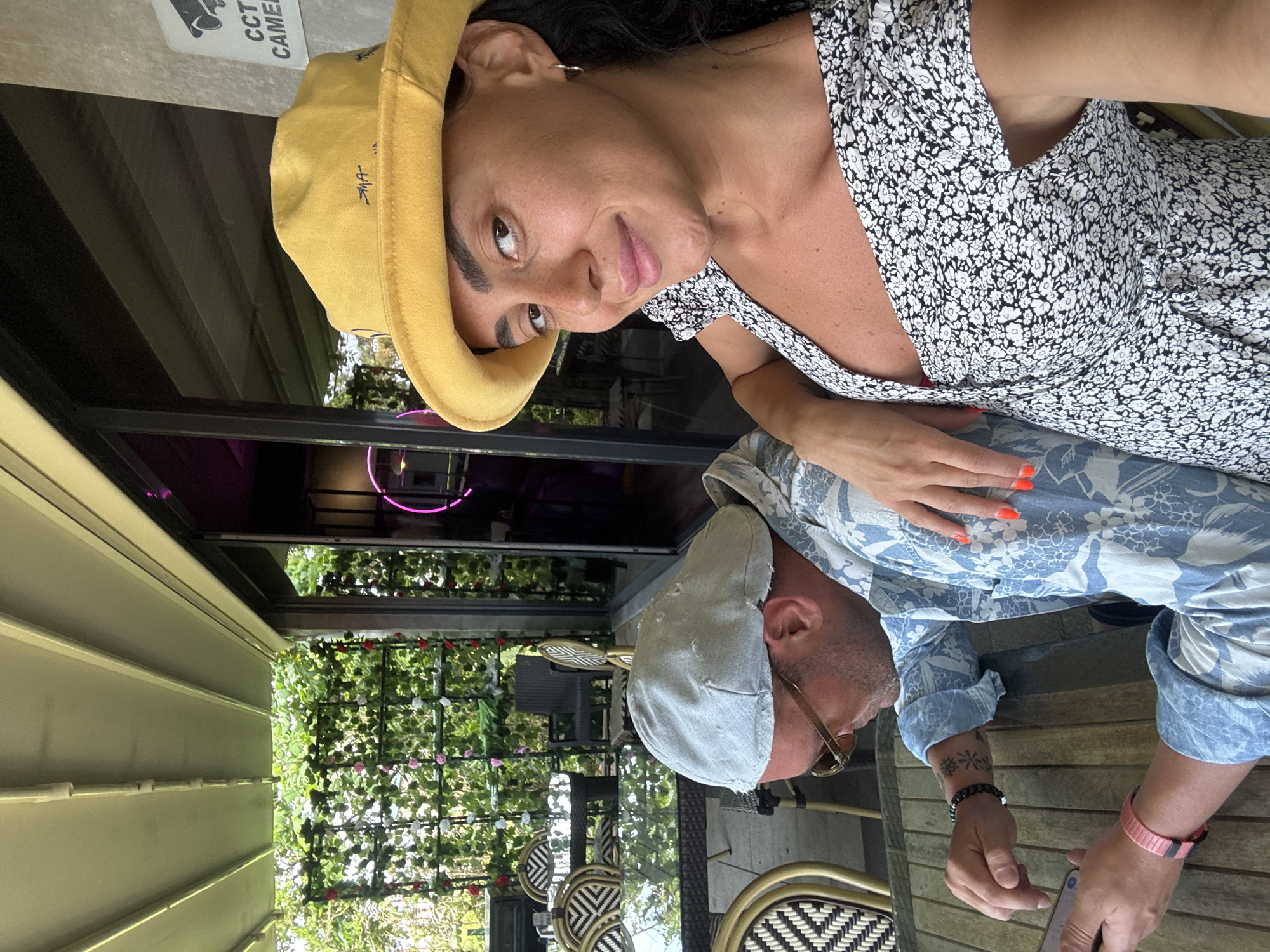 A man and a woman are seated at a table on an outdoor patio. The man, wearing a blue floral shirt and a light grey cap, looks down at his phone. The woman, wearing a yellow hat and a black and white floral dress, smiles at the camera. Behind them, a wall of greenery with pink and red flowers is visible. The scene appears to be in Batumi, Georgia.