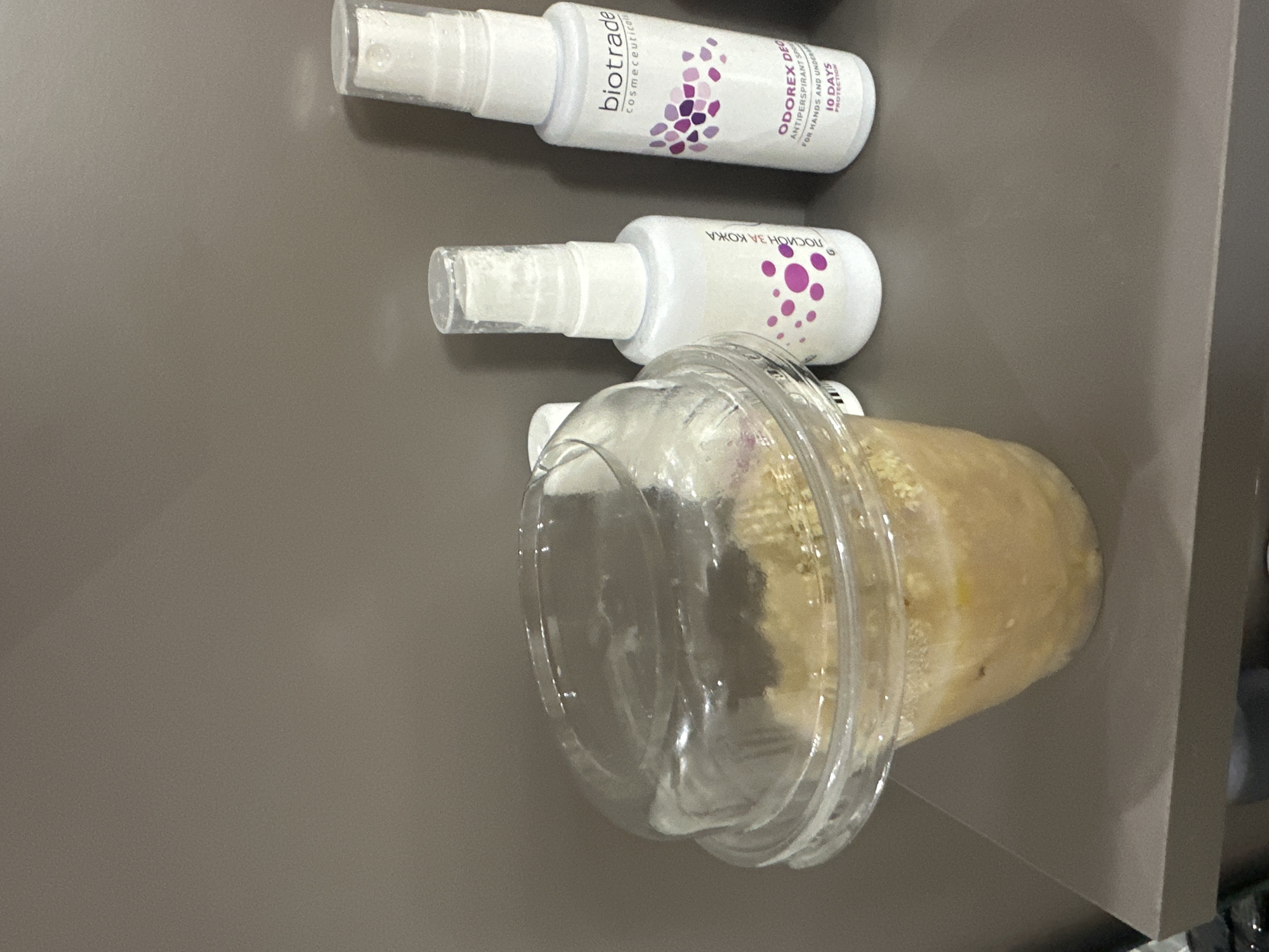 The image displays three items arranged on a flat, plain, medium-brown surface. From top to bottom, there are two white spray bottles, with a clear plastic cup positioned to the right of the bottom bottle.
The top bottle is labeled "biotrade Cosmeceuticals" and features a design with purple and white abstract shapes. The visible text clearly identifies the product as "ODOREX DEO ANTIPERSPIRANT SPRAY FOR HANDS AND UNDERARMS 10 DAYS PROTECTION". It has a clear plastic spray cap.
The second white spray bottle, situated below the first, also has a purple dotted design. The text on this bottle is partially visible and appears inverted, with the words "ЗА КОЖА" (for skin) and what looks like "носно" decipherable. It also features a clear plastic spray cap.
To the right of the second bottle, a clear plastic cup contains a light-colored, yellowish, and somewhat textured substance, possibly a blended food item like a smoothie or oatmeal. The cup has a transparent domed lid that is slightly ajar.
The background is a uniform, unadorned surface, which darkens slightly towards the right edge, possibly indicating a shadow or the inside of a container. The image captures a static arrangement of personal care products and a beverage or food item. There is no discernible activity, specific time of day, or location cues beyond an indoor setting.