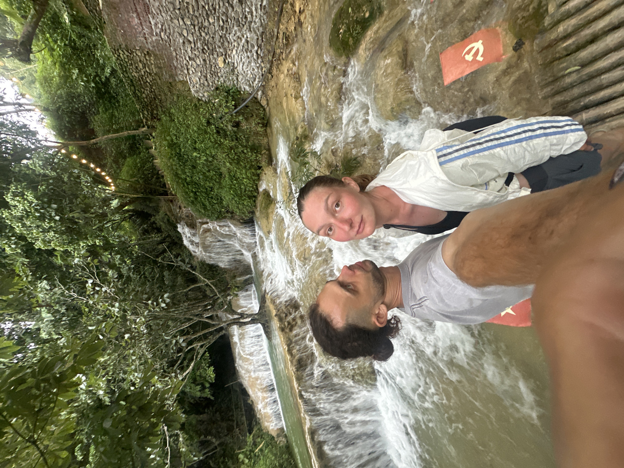In a selfie taken at the Kuang Si Falls in Cổ Lũng Commune, Vietnam, a couple stands before cascading turquoise waters. The man, positioned on the left, has dark curly hair pulled back in a bun and is looking off-camera. He wears a grey t-shirt. The woman, on the right, has light brown hair and is looking at the camera. She is wearing a black tank top and a white jacket with blue stripes. The background features lush green foliage and a multi-tiered waterfall. A small red flag with a yellow hammer and sickle symbol is visible on the rocks to the right of the couple. The overall scene is one of natural beauty with a touch of political iconography.