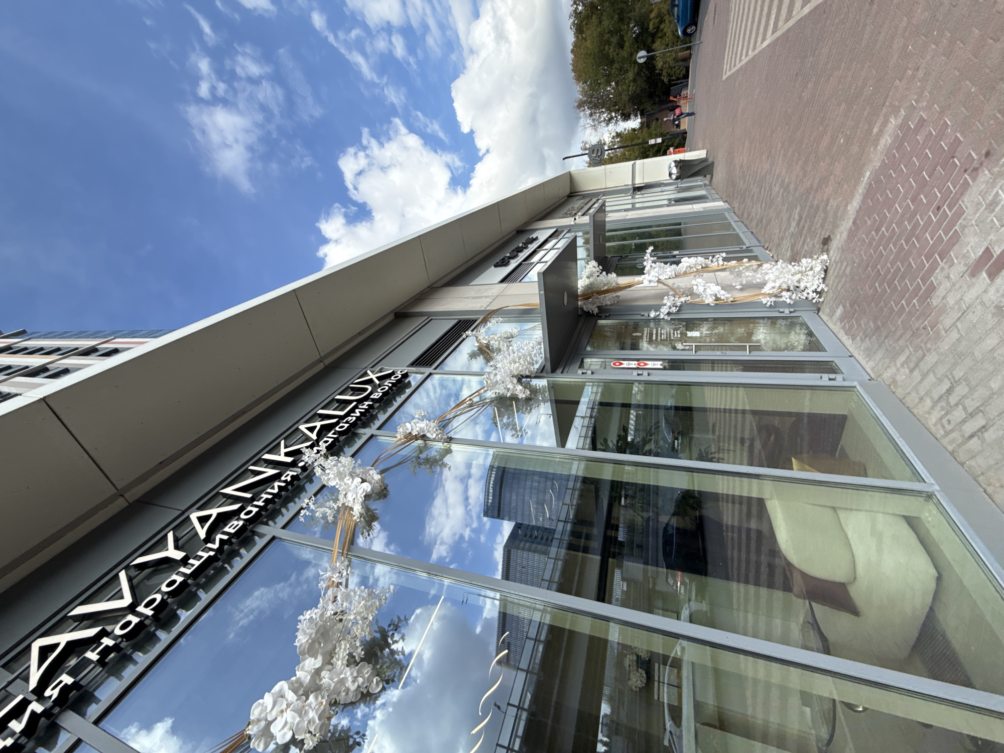 The image shows a storefront with large glass windows on a bright, sunny day with scattered clouds. The building appears modern with a grey facade and a cantilevered section. The storefront has signage in white lettering that reads "AVVYANKALUX" and further text in Russian which appears to be a business name related to hair extensions or a salon. Decorative white floral arrangements are placed on the exterior of the storefront.
Reflected in the glass windows are a highway overpass, several modern buildings, and the sky. Inside the store, a comfortable seating area with a light-colored sofa and pillows is visible through the glass.
To the right of the storefront, a brick-paved sidewalk leads away from the building. A few people are walking on the sidewalk in the distance, and a blue car is parked on the right side of the frame. The overall scene suggests a commercial area in an urban environment. The location is identified as Khimki, Russia.