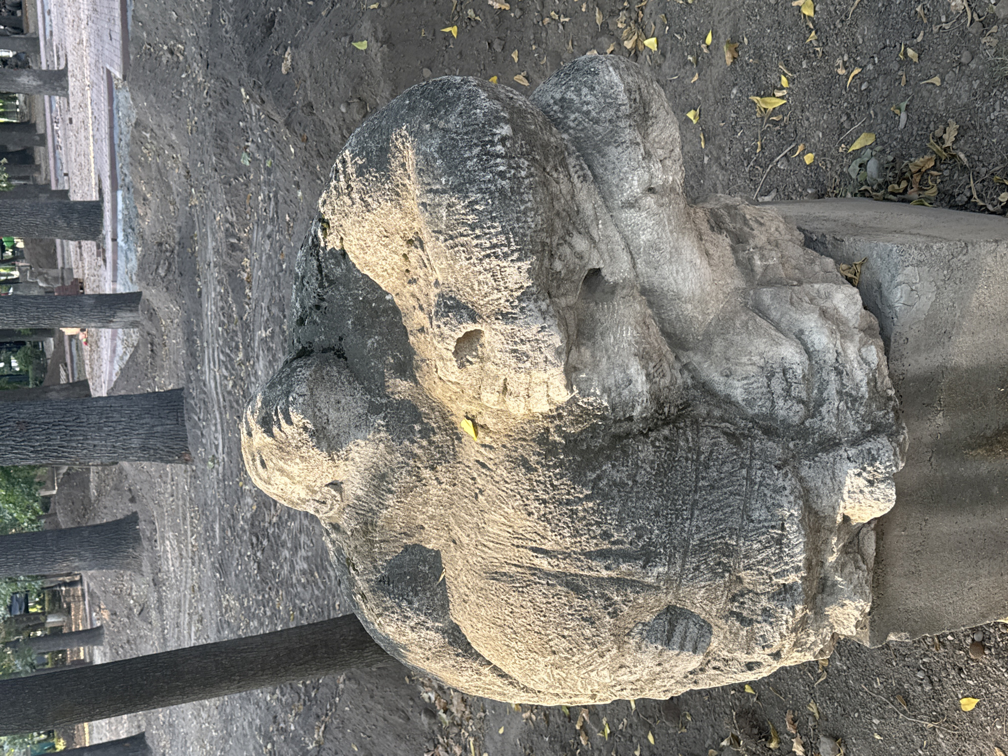 The image shows a stone sculpture of a person in the city of Bishkek, Kyrgyzstan. The sculpture is grey and rough, with visible chisel marks. It appears to depict a person with their head tilted back and their arms wrapped around themselves. The sculpture is placed on a concrete pedestal in an outdoor setting. The background shows trees and a path, suggesting a park or public area. The ground is covered with dirt, fallen leaves, and small stones. There are piles of dirt in the background, indicating some construction or landscaping work. The lighting suggests it is either early morning or late afternoon, with shadows cast by the trees. The sculpture is the primary subject, occupying the center of the frame. The environment is natural and somewhat unkempt, with signs of human activity in the form of the sculpture and the landscaping.