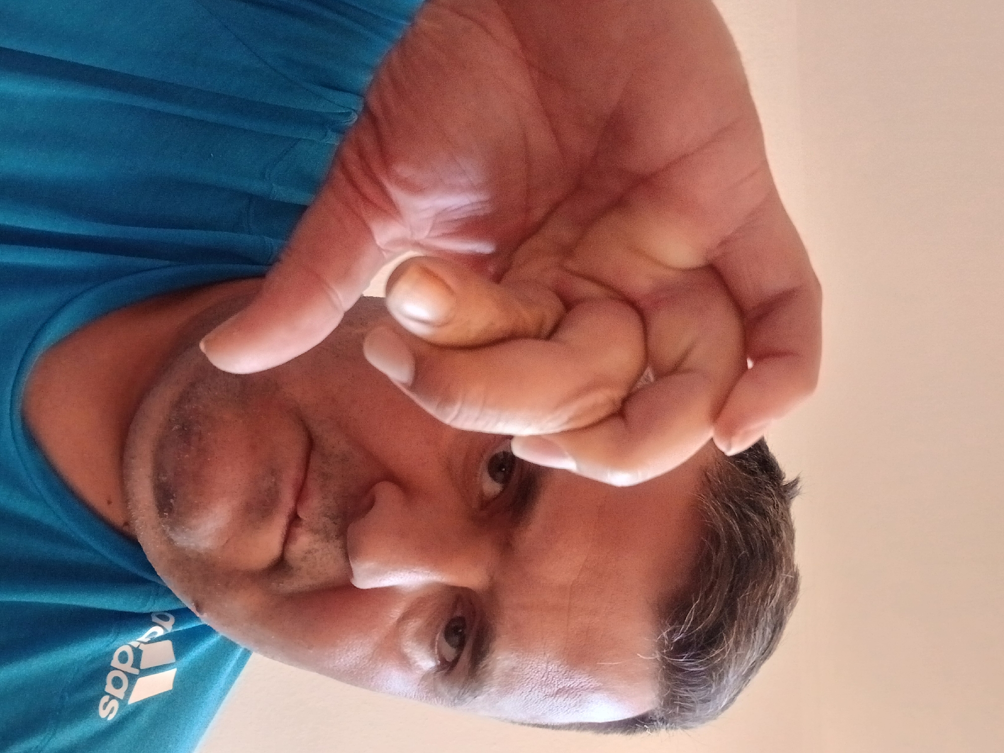A man's face and hand are visible in a close-up selfie. He is wearing a blue t-shirt with the Adidas logo on the right shoulder. The man has short, dark hair with some graying at the temples, and he appears to be in his 40s or 50s. He is looking directly at the camera with a neutral expression. His left hand is raised in front of his face, obscuring part of it, with his fingers curled into a fist. The background is a plain, light-colored wall, suggesting an indoor setting. The lighting is soft and diffused. There is no indication of the specific location of Pfarrkirchen, Germany.