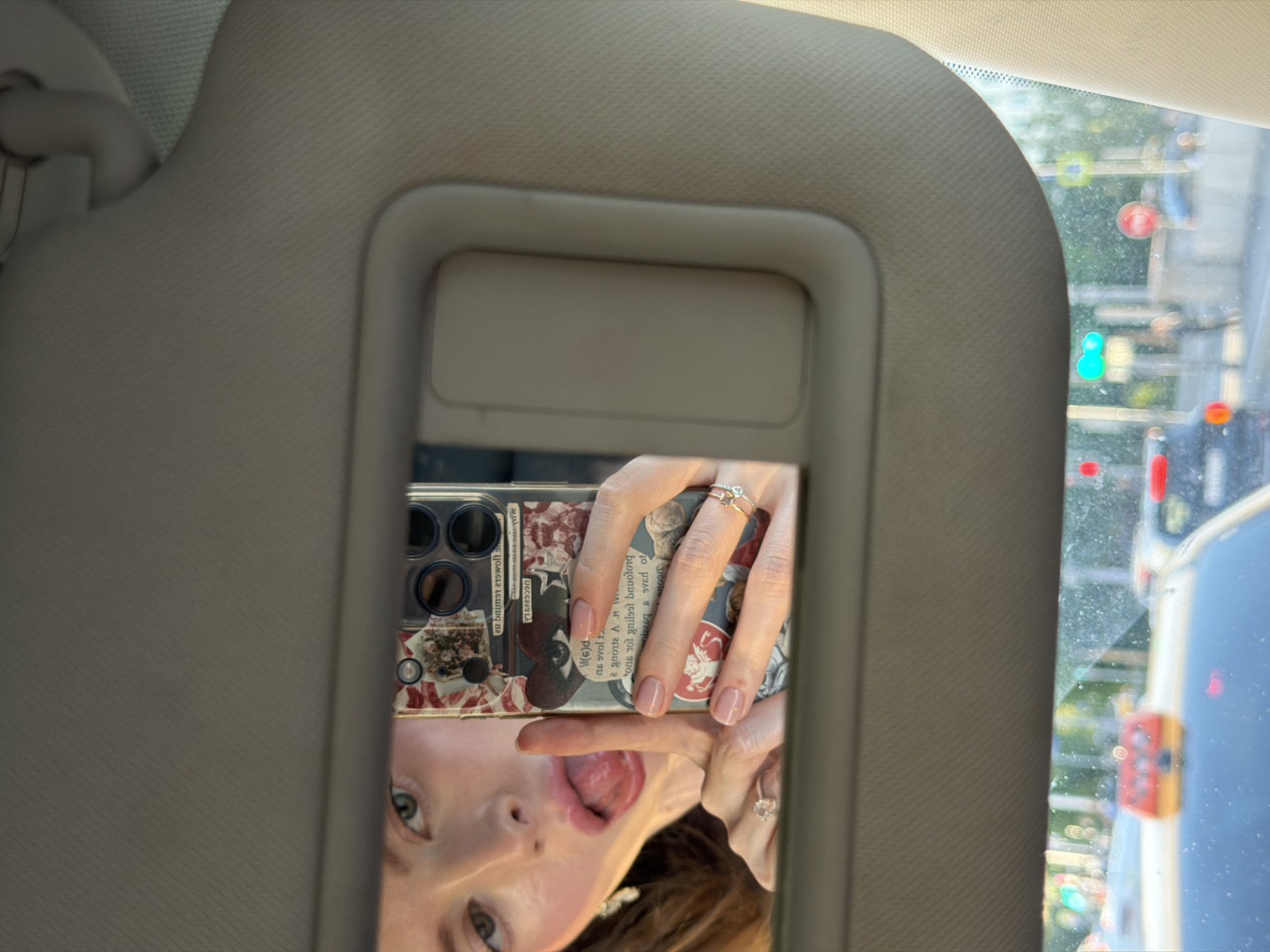 The image displays a playful mirror selfie taken inside a car. The central focus is the reflection of a person's face, appearing upside down, within the rectangular vanity mirror of a beige sun visor. The person has light skin, green eyes, and brown hair, and is playfully sticking out their tongue, indicating a lighthearted mood.
Their right hand, adorned with two rings—one featuring a clear stone and another with multiple thin bands—holds a smartphone. The phone, identifiable by its triple-lens camera module, is fitted with a clear case that showcases decorative inserts. These inserts consist of a vintage-style collage with faded images, possibly flowers, and fragmented, reversed text in an old-fashioned font. An example of the mirrored text reads "20 smowolo sduilod" which, when deciphered, suggests "holding flowers 02".
Through the car's front window, an out-of-focus urban environment is visible. This includes blurred green trees, vibrant red and green traffic lights, and indistinct outlines of buildings or other vehicles, suggesting a city street in Moscow, Russia, as per the provided context. The bright reflections and visible outdoor elements indicate that the photo was taken during the daytime. The overall activity is a casual, self-portrait moment captured while the individual is in a car, likely stationary or moving slowly.