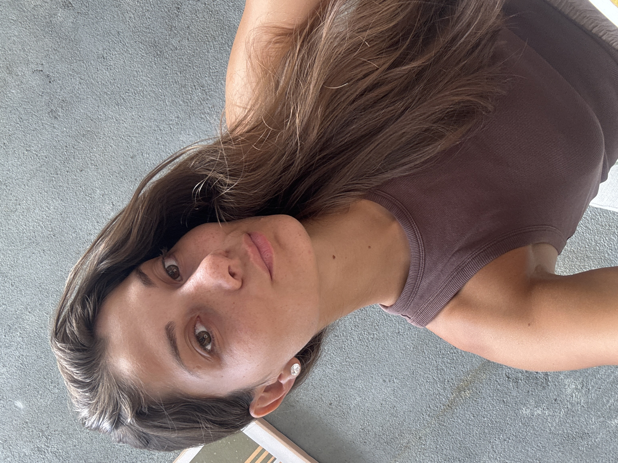 A woman with long brown hair and fair skin is shown in a close-up, low-angle shot. She is wearing a brown sleeveless top. Her face is lightly freckled, and she has brown eyes. She is wearing a silver stud earring in her left ear. The background is a gray concrete surface. There is no indication of the location being in Đà Nẵng, Vietnam.