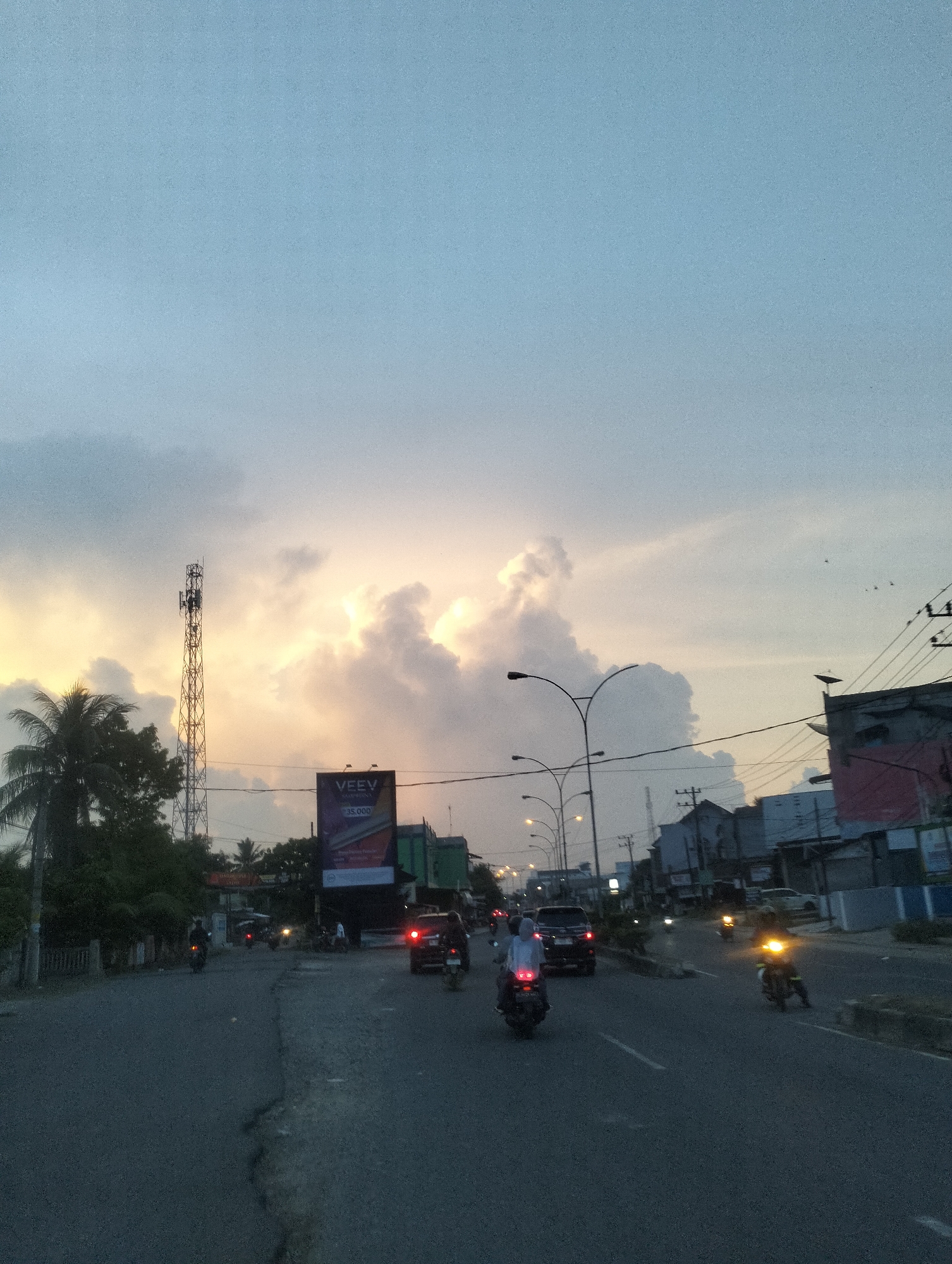 This image captures a bustling street scene in Lhokseumawe, Indonesia, during dusk or early evening. The sky dominates the upper half, featuring a gradient from light blue to warmer golden and orange hues peeking through a dense formation of clouds on the horizon, indicating a setting sun.
The central focus is a multi-lane asphalt road, busy with traffic. Numerous motorcycles and scooters, many with their headlights and taillights illuminated, are traveling in both directions. Several cars or SUVs are also visible amidst the two-wheeled vehicles. Individuals are seen riding the motorcycles, their forms silhouetted or subtly highlighted by ambient and artificial light.
On the left side of the road, a tall, metal lattice telecommunications tower stands against the vibrant sky. Below it, dense green foliage, including palm trees, lines the street. A large, illuminated billboard is prominent, displaying the word "VEEV" and the number "35,000" below it, suggesting an advertisement.
The right side of the image features a line of buildings, appearing to be commercial or residential structures, with overhead utility poles and wires crisscrossing the frame. Streetlights with curved arms are illuminated, casting light onto the roadway. Birds can be seen flying in the distant sky towards the right. The road surface shows signs of wear with some visible cracks. The overall scene conveys an active urban environment as the day transitions to night.