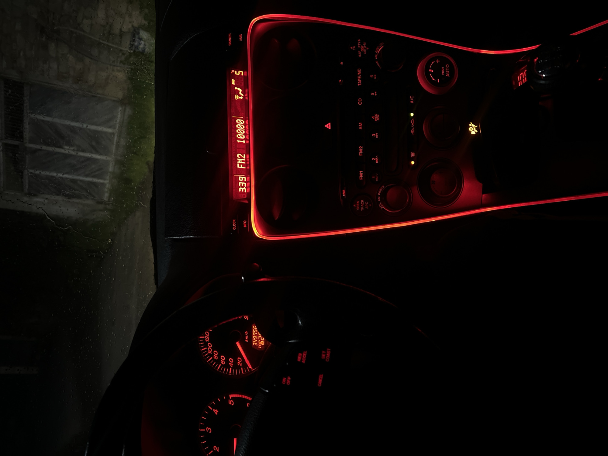 The image depicts the interior of a car, focusing on the dashboard and steering wheel. The lighting is predominantly red, illuminating the car's instruments and controls. The radio display shows "3:39 FM2 10000." The instrument panel displays a speed of approximately 15 km/h and an odometer reading of 349756. The steering wheel includes cruise control buttons. Outside the car window, a dimly lit building or structure is visible through a rain-streaked window. There is no information to determine the location as the city of Kribul, Bulgaria.