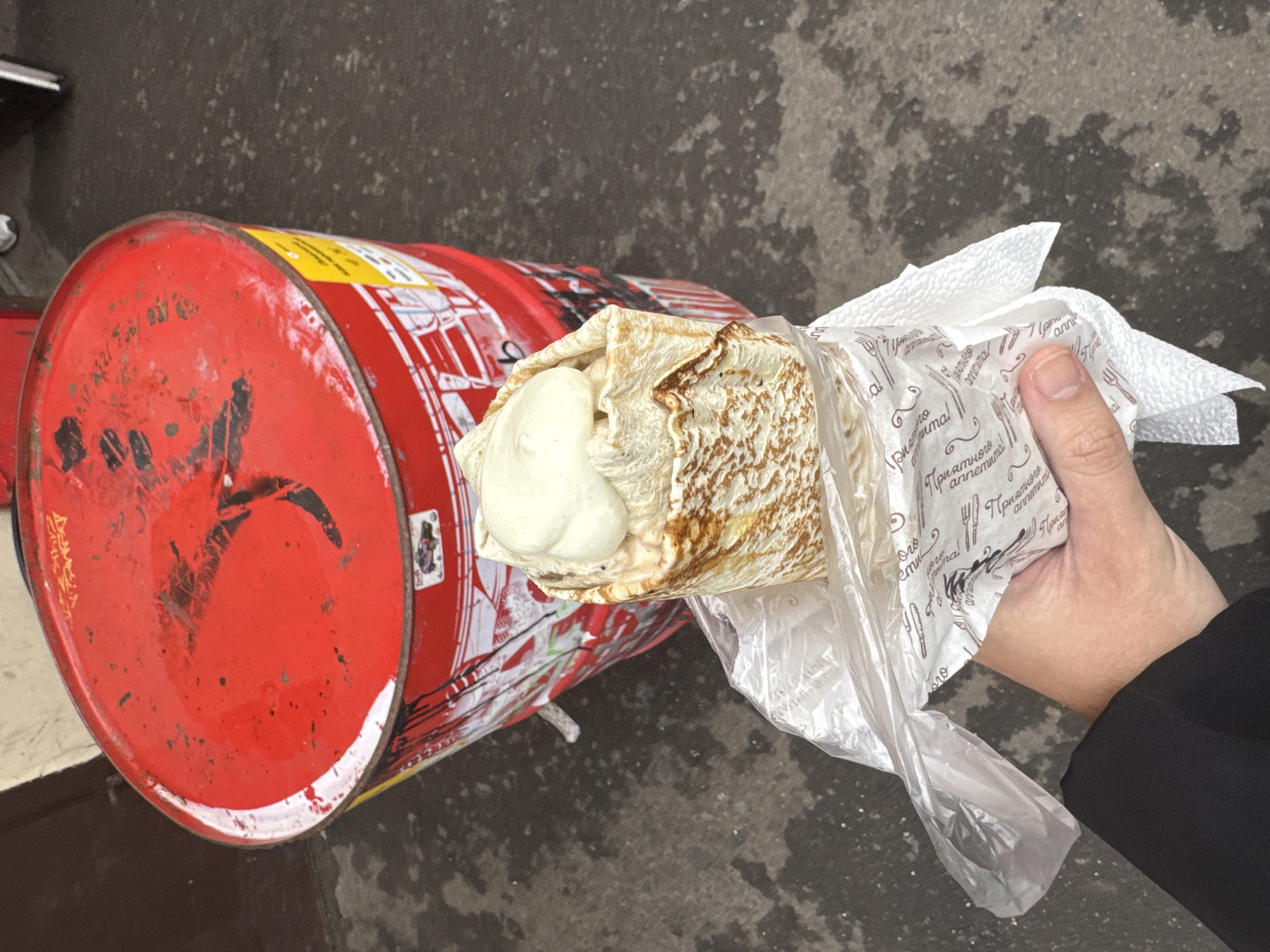 The image shows a hand holding a shawarma wrapped in white paper and a clear plastic bag. The shawarma is placed on a red metal barrel with black markings. The background is a concrete surface, likely a sidewalk. The "Приятного аппетита!" text on the paper suggests a location where Russian is spoken, possibly Moscow, Russia. The person holding the shawarma is wearing a black jacket.