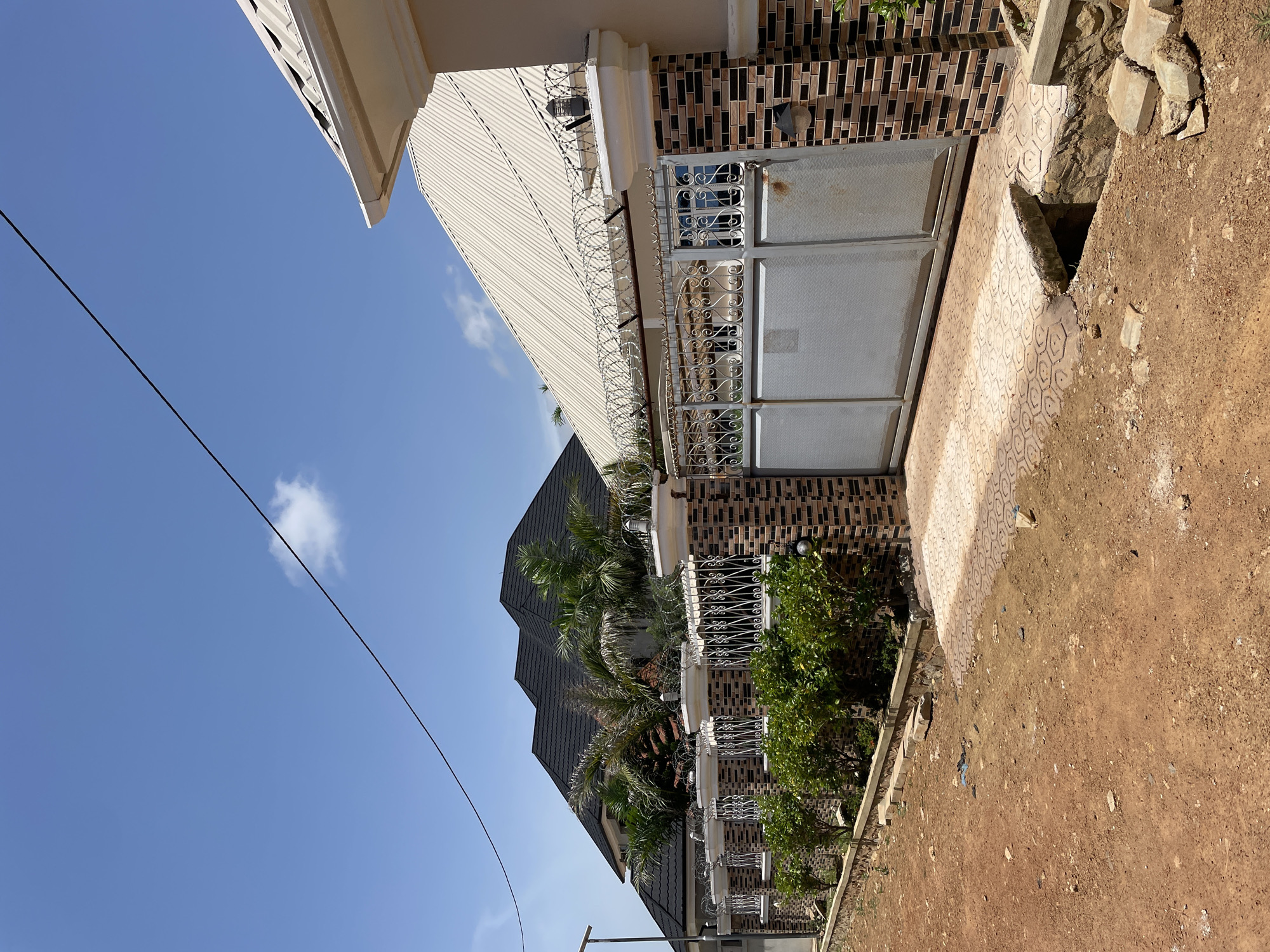 This is a medium-angle, eye-level, exterior shot of a residential property in Rayfield, Nigeria, on a sunny day. The foreground is a rough dirt road with some scattered debris. Leading into the property is a paved driveway with a hexagonal tile pattern. The driveway leads to a large, corrugated metal gate. To the right of the gate is a brick wall with brown and black bricks, and a decorative concrete cap. Above the gate and wall is barbed wire fencing. Behind the gate and fence, the property is lined with neatly trimmed green bushes. Further back, a large house with a dark, tiled roof is visible, partially obscured by palm trees. To the right of this house is another house with a cream-colored, corrugated metal roof, and a beige exterior wall. The sky is blue with a few wispy white clouds, and a power line runs diagonally across the upper portion of the image.