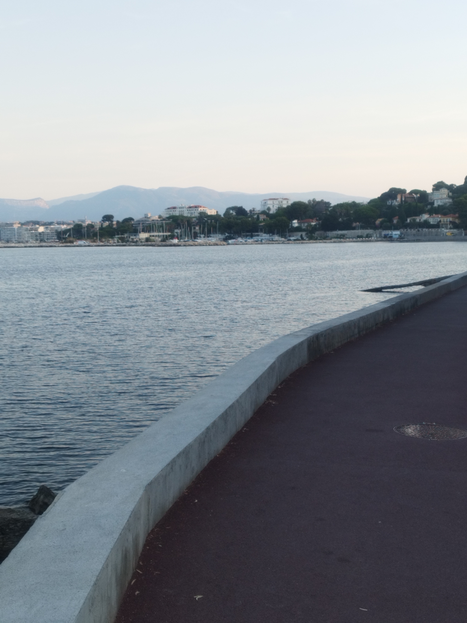 An outdoor, vertical, medium shot captures a paved walkway bordering a body of water in Antibes, France. The walkway is constructed of dark, reddish-brown asphalt and a raised, light gray concrete barrier. A circular manhole cover is visible on the asphalt surface to the right of the frame. The water is a calm, deep blue, with subtle ripples reflecting the overcast sky. In the distance, a coastline is lined with buildings and trees, backed by rolling hills that fade into a hazy, pale sky. The scene is devoid of people.
