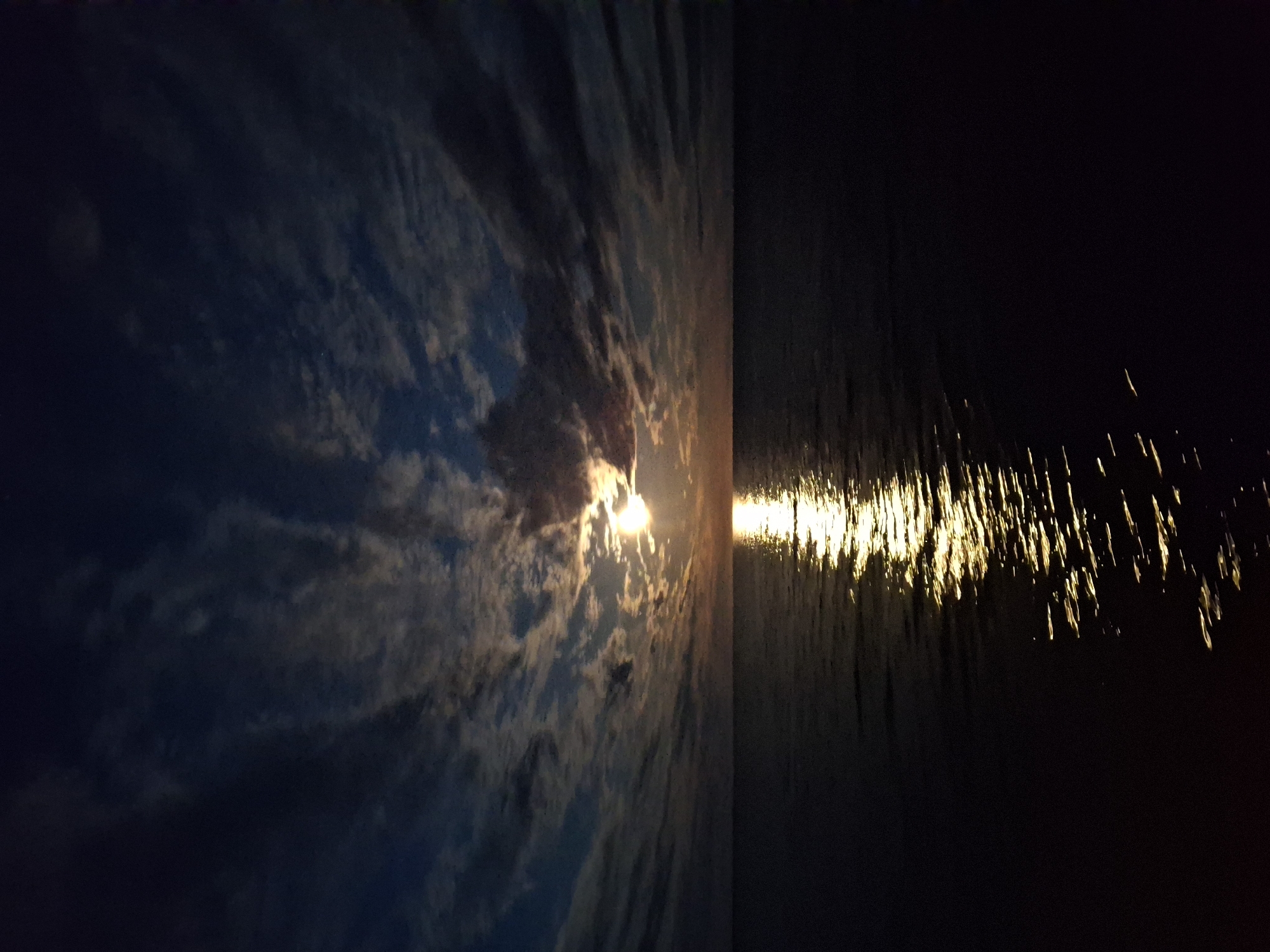 The image depicts a dark seascape at night. On the left, a cloudy sky with a bright light source, possibly the moon or a distant light, partially obscured by clouds. The right half of the image shows the dark water with light reflecting on the surface, creating a series of bright streaks. The scene is likely near a body of water in an outdoor environment. The lack of identifiable landmarks makes it impossible to determine the specific location in the city of Unknown, Latvia. There are no people or specific objects visible other than the water and the cloudy sky.