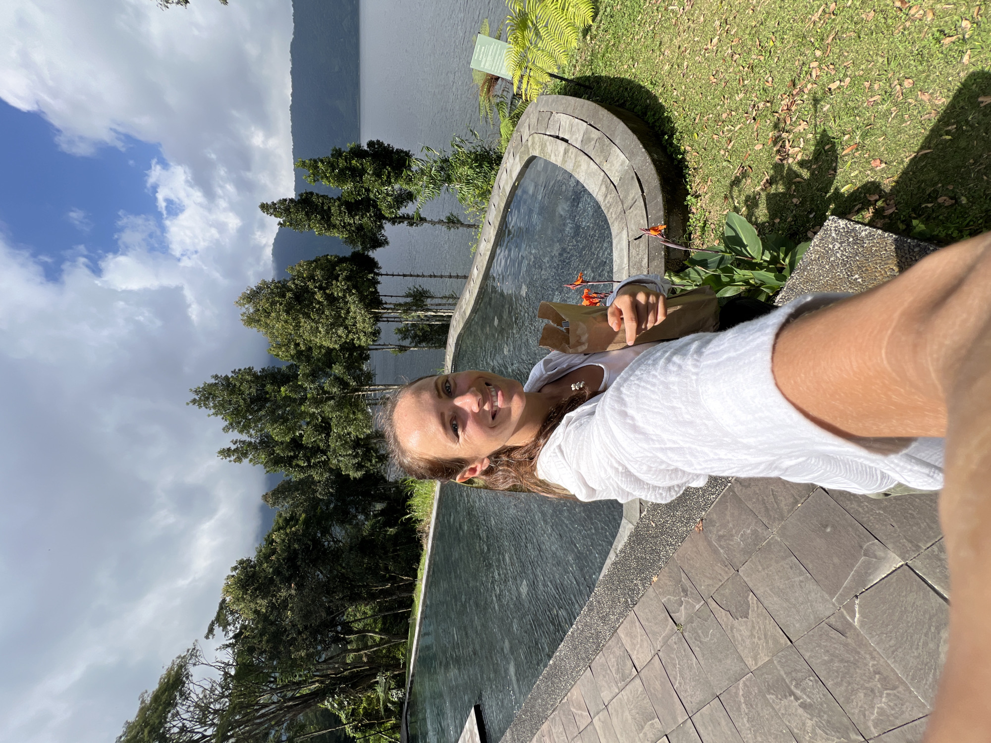 A woman with shoulder-length brown hair, wearing a white hooded jacket and a white top, is taking a selfie in front of a dark swimming pool with a stone border. She is smiling and holding a brown paper bag in her right hand. Behind her, tall trees line the edge of the pool, and a large body of water, likely Lake Batur, is visible in the distance, with mountains and clouds in the background. The area is surrounded by lush green grass and stone paving. The location is identified as Baturiti, Indonesia, suggesting a scenic resort or natural setting.