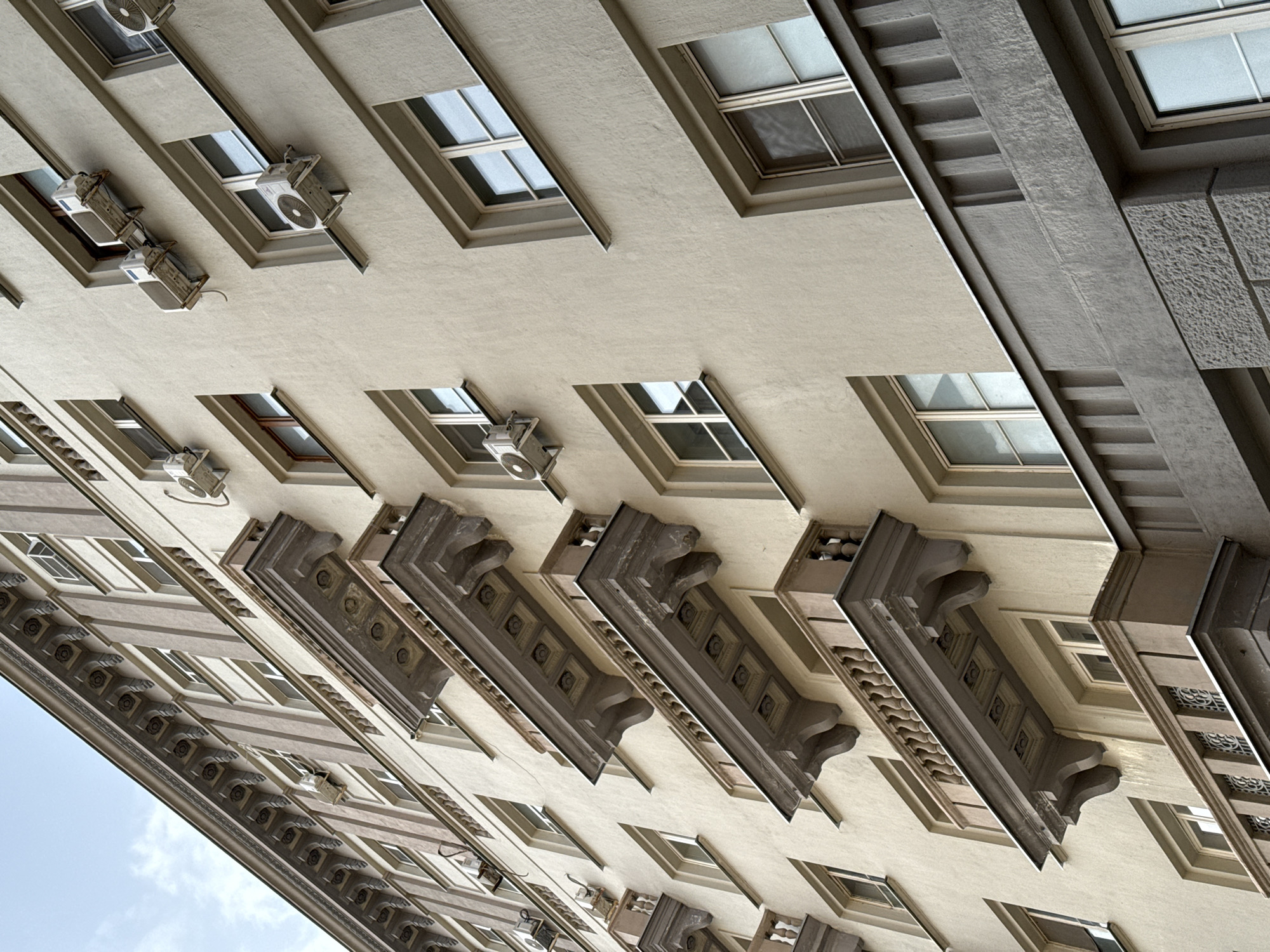The image displays an upward-angled view of the facade of a multi-story residential building located at Bolshaya Polyanka Street, 1/3, in Moscow, Russia. The architecture presents a Stalinist Empire style, characterized by a light beige stucco finish contrasting with darker brown-grey decorative elements. These elements include a prominent, elaborately carved cornice at the top, vertical pilasters, and several projecting balconies. The balconies feature balustrades with classical balusters or rectangular vertical elements, dark grey top railings, and undersides decorated with recessed square panels containing circular motifs. Numerous white, external air conditioning condenser units, some exhibiting rust and visible piping, are irregularly affixed beneath or adjacent to the rectangular, multi-paned windows on various floors. The lowest visible section of the building features a dark grey, textured stone facade with simple vertical balustrades. The sky, showing patches of light blue with sparse white clouds, is visible in the upper left corner. No individuals or discernible activities are present in the scene.