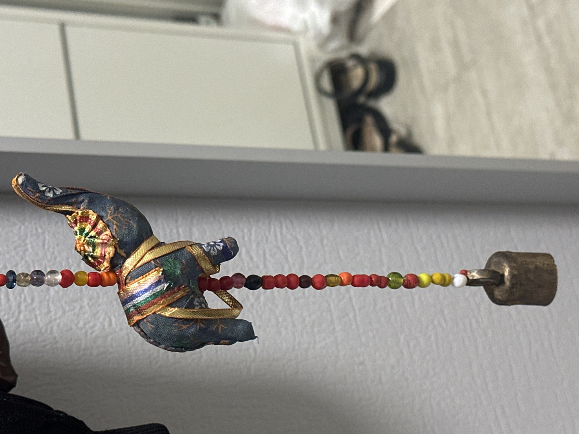 The image shows a close-up of a decorative hanging ornament. The ornament features a small, fabric-covered elephant with colorful striped fabric and gold trim. It is suspended from a string of colorful beads, ranging in color from red and orange to yellow, green, and blue. A small, brass bell hangs at the bottom of the beaded string. The background is blurred, but appears to be an indoor setting with a light-colored wall on the left and a glimpse of furniture and shoes in the background on the right. The image is a shallow depth of field, focusing sharply on the elephant and beads while blurring the surroundings. There is no indication of Nevinnomyssk, Russia, in the image itself.