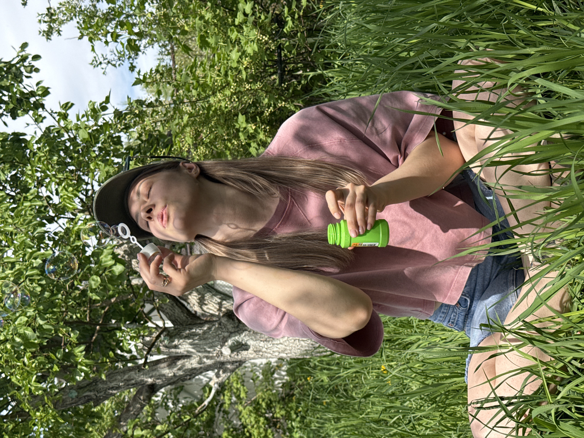 A young woman in a pink t-shirt, denim shorts, and a dark cap is kneeling in tall grass, blowing bubbles. She holds a green bottle of bubble solution in one hand and a bubble wand in the other. Bubbles float near her face, with a tree trunk and foliage behind. The image was likely taken in Chaikovsky, Russia, based on the query, but there is no other corroborating evidence within the photo.