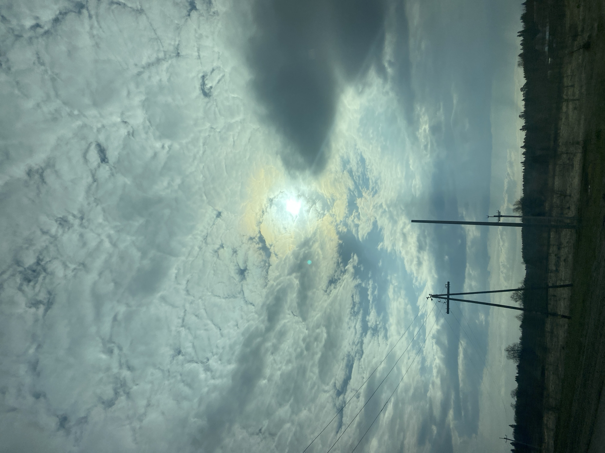 The image shows a daytime scene in what appears to be a rural or semi-rural area. The sky is predominantly cloudy, with patches of blue visible. The sun is obscured by the clouds but still emits a bright glow. Power lines and telephone poles are visible, stretching across the frame, indicating the presence of infrastructure. A treeline or forest is present in the distance. It is not possible to definitively confirm the location as Malaya Bod'ya, Russia, based solely on visual information.