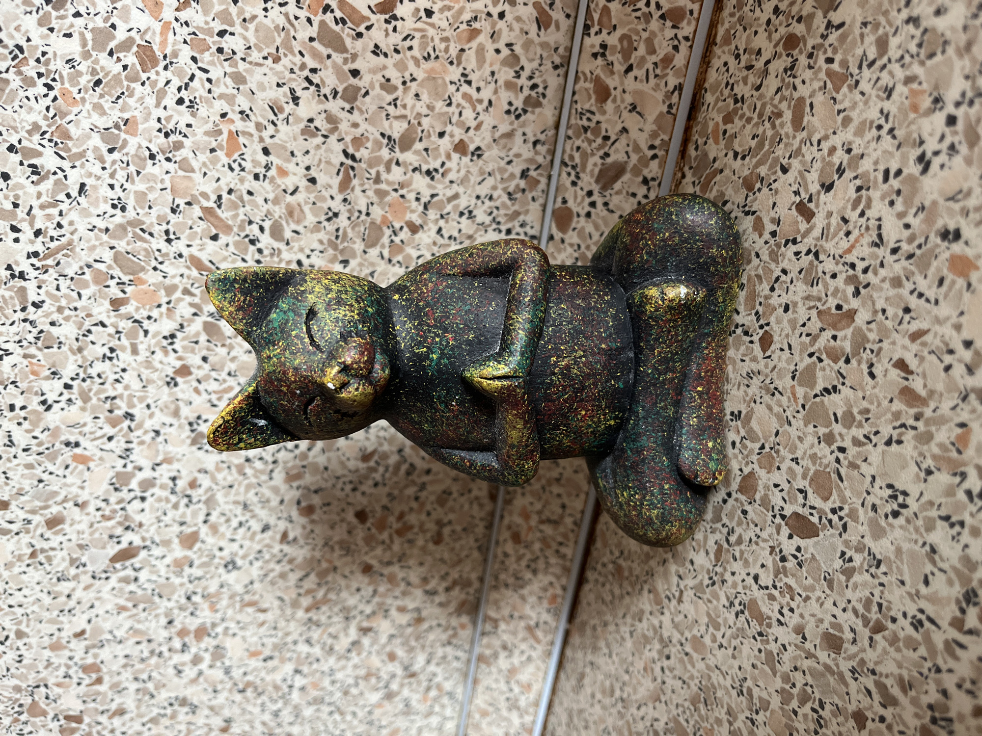 A small, dark statue of a cat in a seated, meditative pose is the central subject of the image. The cat statue is speckled with various colors, including green, yellow, red, and hints of gold, giving it a textured appearance. It has closed eyes and its paws are pressed together in front of its chest. The statue is positioned on a speckled surface that resembles terrazzo, with a lighter background also featuring a similar speckled pattern. Two thin, horizontal metal rods are visible behind the statue. The lighting suggests an indoor environment. The image does not contain any discernible information about the city of Saratov, Russia, or any specific location within it.