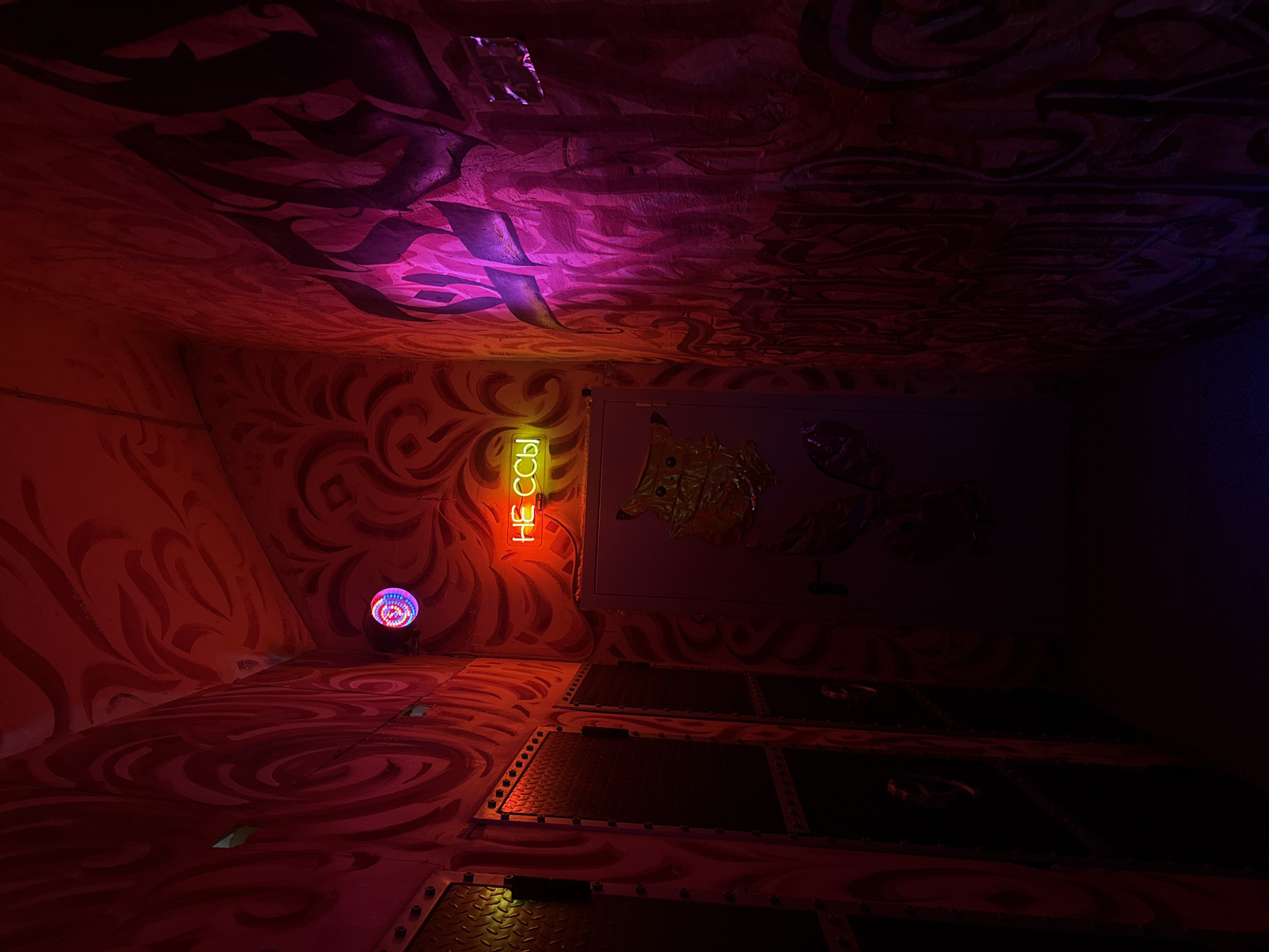 This image shows an interior space in Perm, Russia, with walls and flooring painted with a swirling red and black pattern. A neon sign saying "Не ссы" (Don't Pee) in Russian hangs on one wall. Another wall features a white panel with foil sculptures. A multi-colored light fixture is mounted near the corner. The floor appears to be constructed of metal plates. The lighting is predominantly red and purple, giving the space a surreal atmosphere.