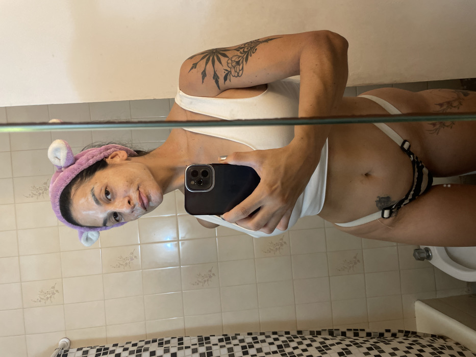 A female subject is depicted taking a mirror selfie in a residential bathroom, located in Buenos Aires, Argentina. The subject, an adult female, is shown from mid-thigh to above the head. Her face is covered with a white, pasty substance, consistent with a facial mask, and she wears a lavender-colored plush headband with white ear-like protrusions. She is clad in a white sleeveless crop top and a two-toned (white and dark-striped) thong-style undergarment with visible strap rings. Tattoos are present on her left upper arm/shoulder (a dark, radiating plant or floral design), her lower abdomen (an indistinct dark mark to the left of the navel), and a fainter, lighter design on her inner left thigh. She holds a dark-colored smartphone, featuring a prominent camera array, in her right hand, which has metallic gold nail polish on the thumbnail. The bathroom environment includes light-colored square wall tiles, some with faint floral patterns. A black and white checkered shower curtain is partially visible on the far left, and a white porcelain toilet or bidet is visible in the lower left background. A vertical reflective strip, consistent with a mirror's edge, runs down the right side of the image.