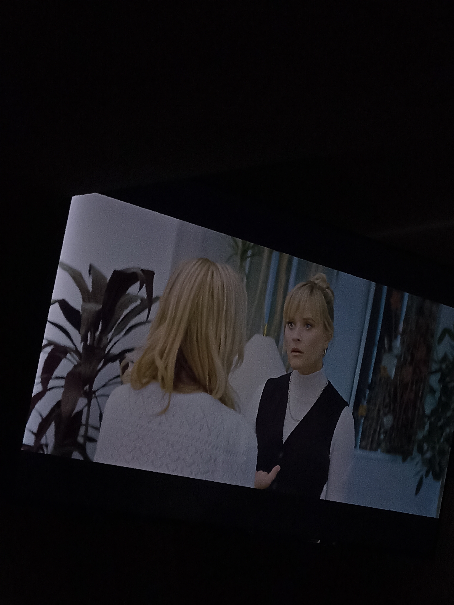 The image shows a television screen in a dark room. On the screen, two women are visible in an interior scene. The woman on the left has her back to the viewer, with blonde hair and wearing a white sweater. The woman on the right, identified as Reese Witherspoon, is wearing a white turtleneck and a black vest. She has her blonde hair in an updo and appears to be in a state of mild surprise or concern. The background includes plants and what seems to be a window or a curtain. The location of San Bernardino Tlaxcalancingo, Mexico, is not depicted in the image and there is no evidence linking the image to that location.
