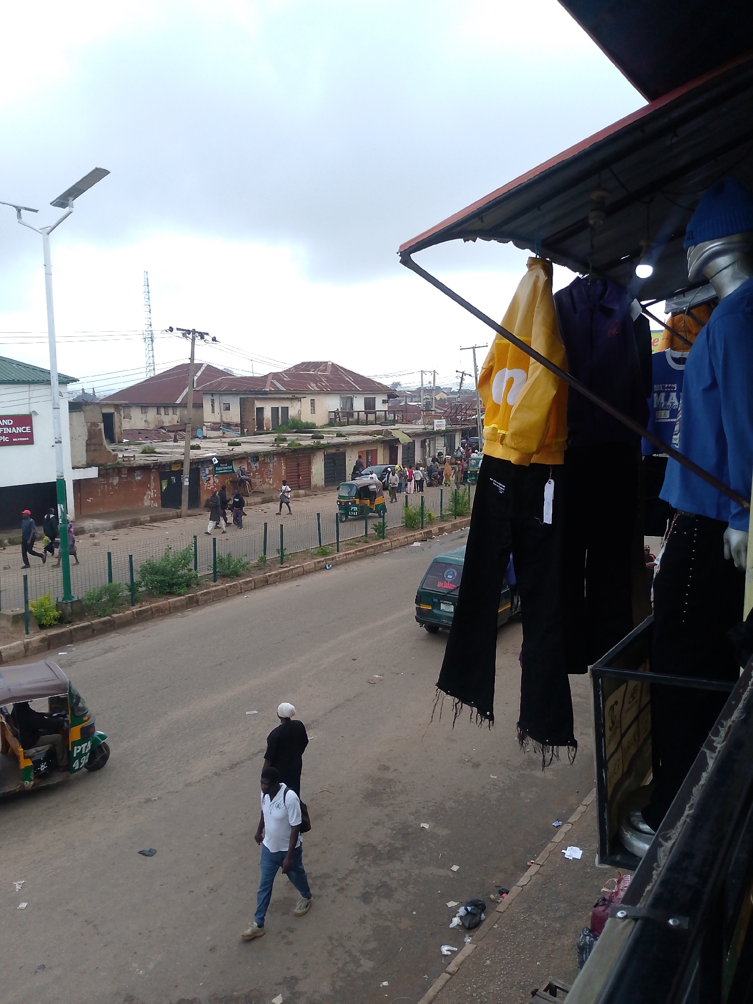 The image depicts a street scene in Bukuru, Nigeria, captured from an elevated perspective, likely a balcony or upper floor. The street is paved and appears busy, with several people walking along the sidewalk and crossing the road. A green and yellow auto-rickshaw, a common mode of transport in Nigeria, is visible on the right side of the street, with a driver inside and "PTA 497" visible on its side.
In the foreground, clothing is displayed on mannequins or racks, suggesting a shop or market stall. A yellow jacket with white lettering and black pants hang prominently. Further back, buildings line the street, some appearing to be older structures with corrugated iron roofs and weathered walls. Utility poles and electrical wires crisscross the sky, typical of urban infrastructure.
The sky is overcast, indicating a cloudy or possibly rainy day. A white solar-powered street lamp stands on the left side of the image, its panel angled towards the sky. The overall impression is one of a bustling urban environment with everyday activities taking place.