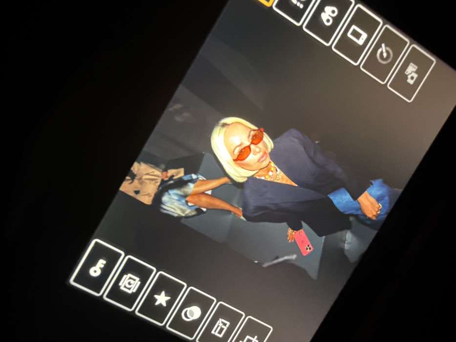 A tilted photograph captures a digital screen displaying an image of two individuals in an indoor, studio-like environment. The primary subject in the foreground is a light-skinned individual with short, blonde hair, wearing small red oval sunglasses, a dark blazer, and blue trousers. This individual accessorizes with multiple colorful beaded necklaces and holds a bright pink smartphone in their left hand, angled downwards, while looking slightly down and to their right.
Behind and to the right of the primary subject, a second individual is partially visible. Their lower body is dressed in a light-colored skirt or dress with a blue and white pattern, paired with light shoes. The upper body and head of this individual are obscured by a beige, crumpled object, possibly a bag, with dark markings on it.
The background within the displayed image is dark and appears to be a featureless or subtly textured wall, indicating an artificial or studio setting with focused lighting on the subjects. The digital screen itself shows portions of a user interface with white outlined icons against a dark gray background on both the left and right sides. Left-side icons include "On," a camera, a star, and a circular symbol. Right-side icons include binoculars, a smartphone, a timer/clock, and a video camera/gallery icon. The specific location within Dubai, United Arab Emirates, is not discernible from the visual information provided.