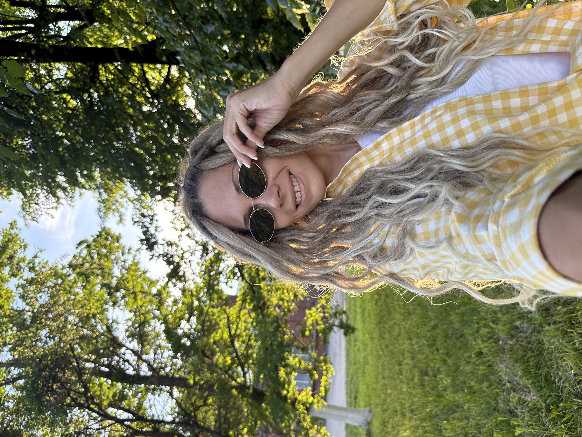 A woman is captured in a medium close-up selfie outdoors, with a backdrop of trees and grass. She is wearing round sunglasses, a white tank top, and an open yellow and white checkered shirt. Her long, blonde, wavy hair is partly visible, along with a few braids intertwined. She is smiling and touching her sunglasses with her right hand. The lighting suggests it is either sunrise or sunset, casting a warm glow on her face and hair. The environment appears to be a park or a yard, with green grass and lush trees. There is a building with red brick visible in the background to the left, partially obscured by trees. The location is specified as Shebunino, Russia.