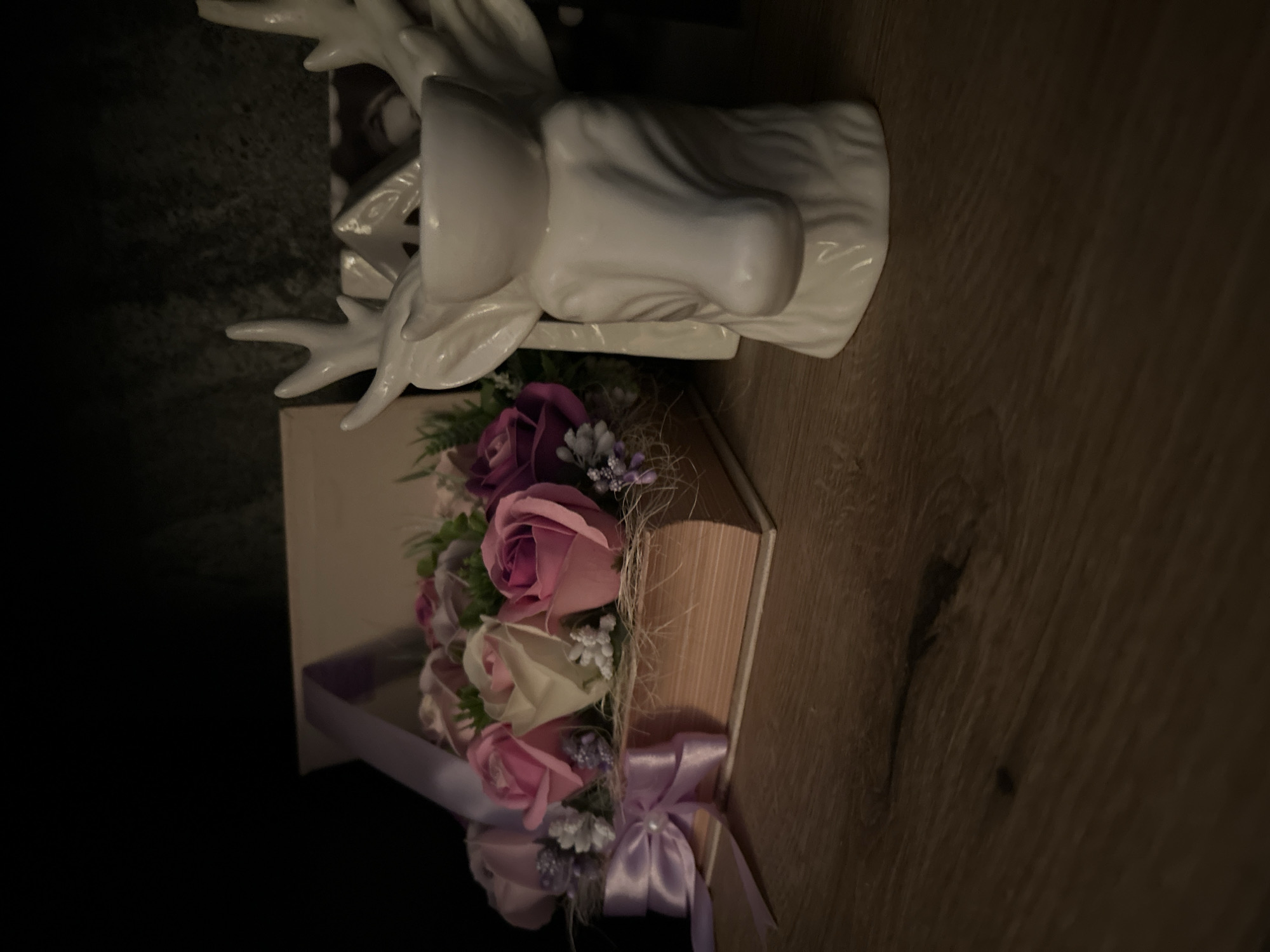 The image shows a close-up view of a decorative arrangement on a wooden surface, likely a shelf or table. The foreground is dominated by a white ceramic figurine shaped like a deer's head with antlers, which appears to be a candle holder. To the left, a box filled with artificial roses in shades of pink and purple is placed on top of what looks like a book. The box is adorned with a pale purple ribbon tied into a bow. The background is dark and out of focus, suggesting an indoor setting with low lighting. There are no people visible in the image, nor are there any specific landmarks that would place the scene in Sofia, Bulgaria.