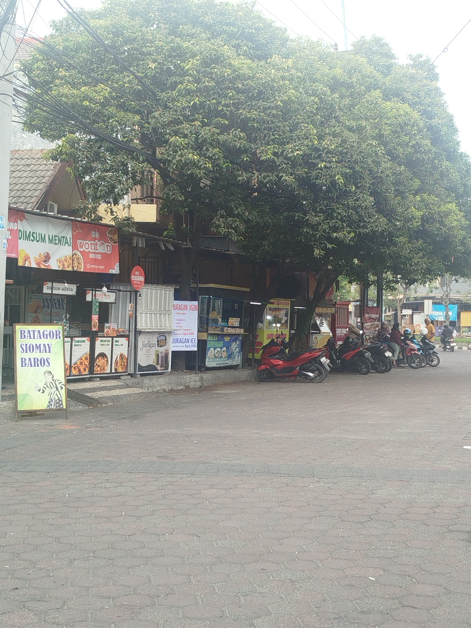 The image shows a street scene in Yosowilangun, Indonesia, with a row of food stalls and parked motorcycles. A large tree with dense foliage dominates the upper portion of the frame. In the foreground, a paved sidewalk is lined with various food vendors. One prominent stall features a sign that reads "BATAGOR SIOMAY BAROS" with a cartoon of a person giving a thumbs-up. Other stalls display menus and images of food, including "DIMSUM MENTAI," "wonton," and "JURAGAN JIGOR." Several motorcycles, including a red one, are parked along the sidewalk in front of the stalls. A few people are visible, some standing near the stalls and one person on a motorcycle in the background. The environment appears to be a commercial area with overhead electrical wires and a building with a tiled roof visible behind the stalls. The overall scene depicts a bustling street-side dining area.