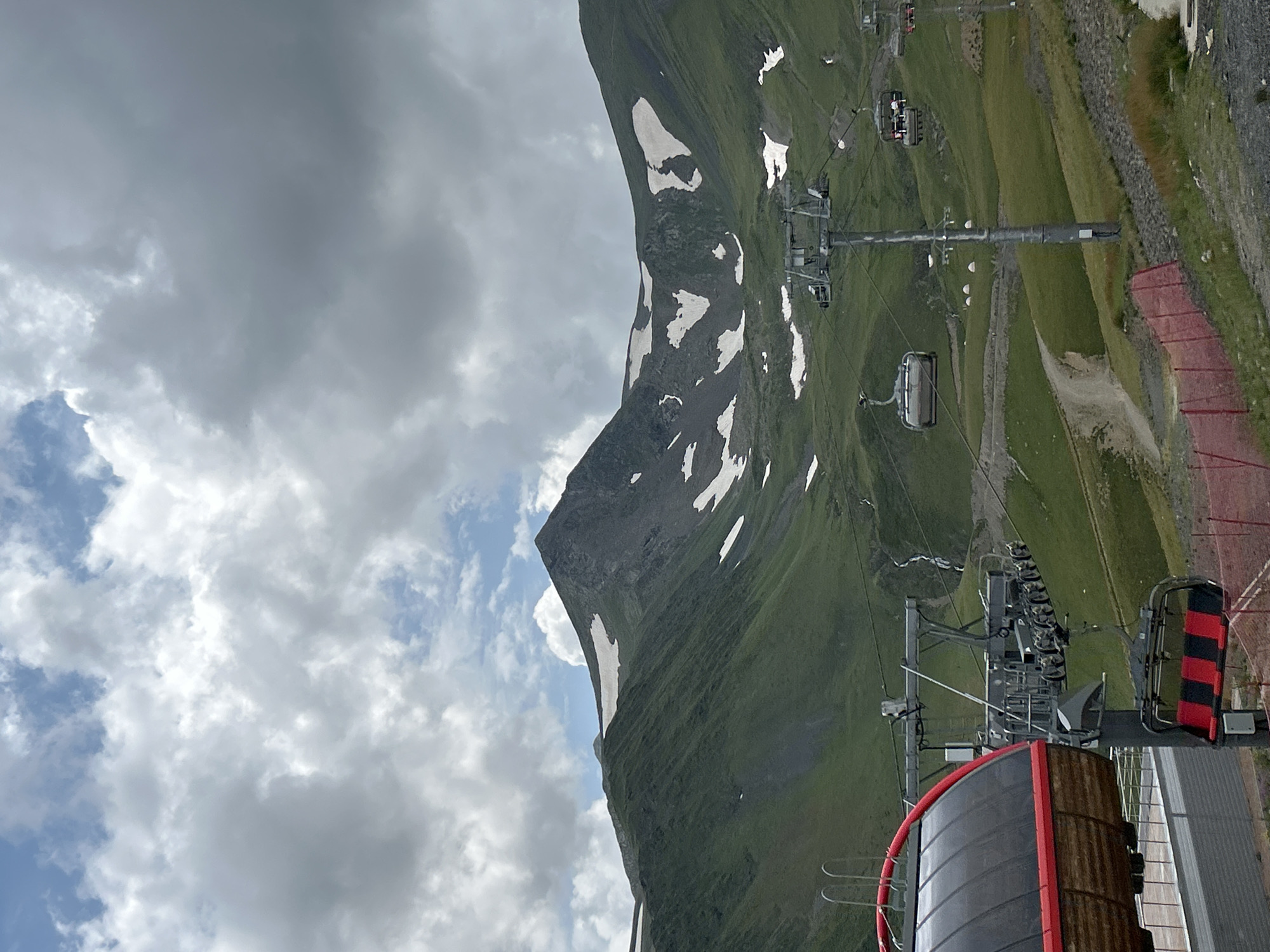 The image depicts a cable car station and mountainous landscape in Lunnaya Polyana, Russia. In the foreground, a red and black cable car cabin is partially visible on the left, with its interior featuring wooden panels and a red trim. Adjacent to it, a ski lift tower supports multiple cable car seats, some of which are red and black striped. The cable car system extends up a grassy mountainside. In the midground, several cable cars are seen traversing the sky above rolling green hills dotted with patches of snow. A dirt path winds through the landscape, and a small waterfall is visible cascading down the mountain. The background is dominated by a rugged mountain peak, with a mixture of green slopes and rocky outcrops, also streaked with snow. The sky above is filled with dramatic, grey and white clouds, with hints of blue sky peeking through. The overall scene conveys a sense of natural beauty and recreational activity in a mountainous setting.