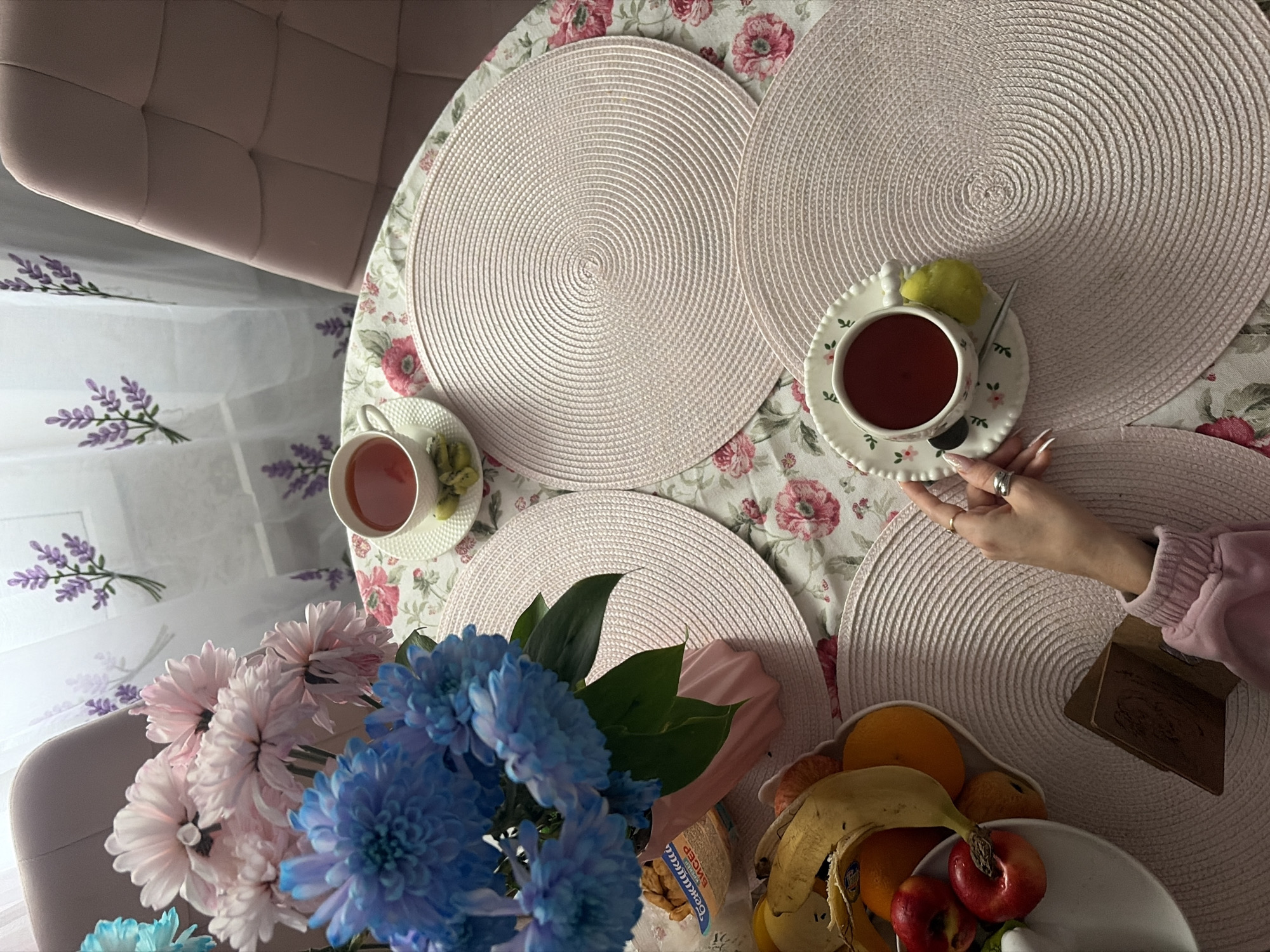 A top-down shot depicts a round table covered with a floral-patterned tablecloth. Four round woven placemats sit on the table. Two teacups on saucers are present, one held by a hand with painted nails wearing a ring. One cup holds tea with a green lemon. A bowl of fruit containing bananas, oranges, and apples is also visible. A bouquet of blue and pink flowers is near the fruit. A portion of a pink chair is visible in the upper left and a second chair in the lower left. A patterned white curtain hangs in the background. There are no definitive clues to confirm that this is Saint Petersburg, Russia.