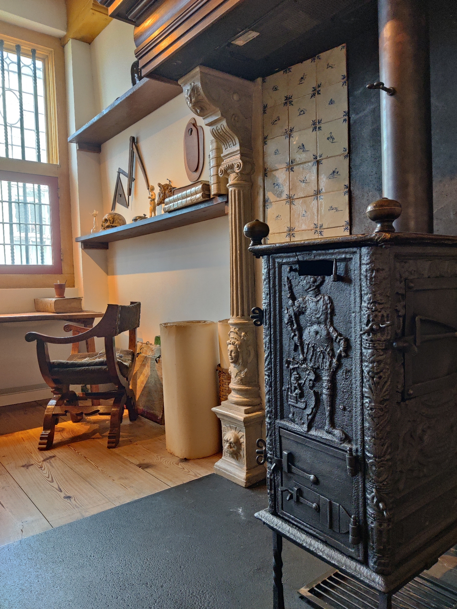 The image depicts a well-preserved, antique interior space, likely a historical residence or museum exhibit in Amsterdam, Netherlands. Dominating the foreground is an ornate, dark metal stove with intricate relief carvings of a knight or soldier. The stove rests on a black metal stand with a grate at the bottom. To the left of the stove, a tall, cylindrical metal pipe extends upwards, suggesting it's a flue. Behind the stove, a wall is partially covered with decorative tiles featuring blue patterns of animals and geometric shapes.
Further to the left, a wooden chair with a dark upholstered seat and back is positioned on a light-colored wooden floor. Next to the chair, a tall, cream-colored cylindrical object, possibly a storage container or a part of an old appliance, stands upright. To the left of the chair and the cylindrical object, a wooden desk or table is visible, with a closed book and a small cup resting on it. A large window with multiple panes and metal bars is situated on the far left, allowing natural light to illuminate the scene.
Above the desk and extending across the wall, two wooden shelves are installed. The lower shelf displays several items, including a skull, a small figurine, a metal contraption resembling a guillotine or a printing press mechanism, and a stack of old books. The upper shelf has fewer items, with a small wooden elephant figurine visible. The wall itself is painted a light, neutral color. The overall atmosphere is one of historical preservation, suggesting a glimpse into a past era within the city of Amsterdam.