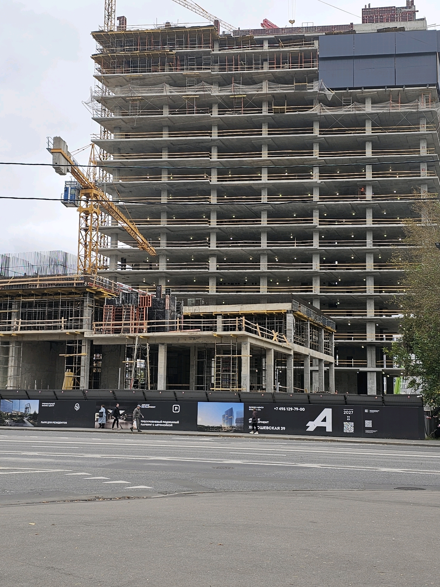 The image shows a multi-story building under construction in Moscow, Russia. The building is made of concrete and steel, with multiple levels of floors and columns visible. A yellow construction crane is positioned to the left of the building, extending over several floors. In the foreground, a black fence with various advertisements and information panels runs along the street. The fence displays images of buildings, text in Russian, and logos. Three people are walking along the sidewalk in front of the fence. The street in front of the fence has white lane markings. The sky is overcast. The construction site appears to be a large urban development project.