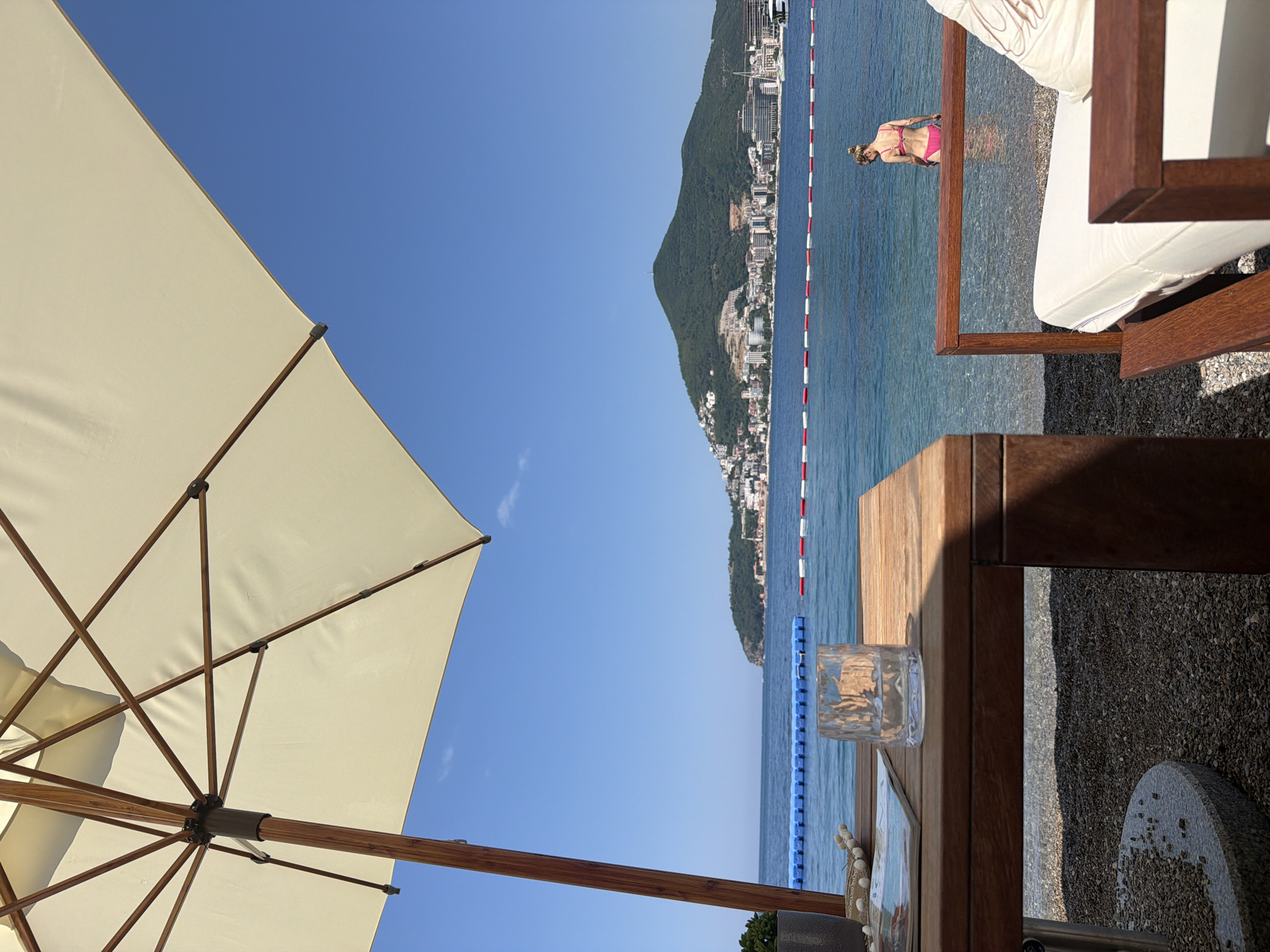 A woman in a pink bikini stands in the shallow, clear blue water of a beach. Behind her, a picturesque coastline is visible, with a densely built city nestled against a lush green mountainside. A red and white buoys mark the edge of the swimming area. In the foreground, a large, cream-colored umbrella provides shade. A wooden table with a glass of water and some decorative shells sits nearby. The scene appears to be set on a sunny day, likely late morning or early afternoon, given the bright light and clear blue sky. The overall atmosphere is one of leisure and relaxation, suggesting a vacation or a day spent at a seaside resort. The location is identified as Budva, Montenegro.