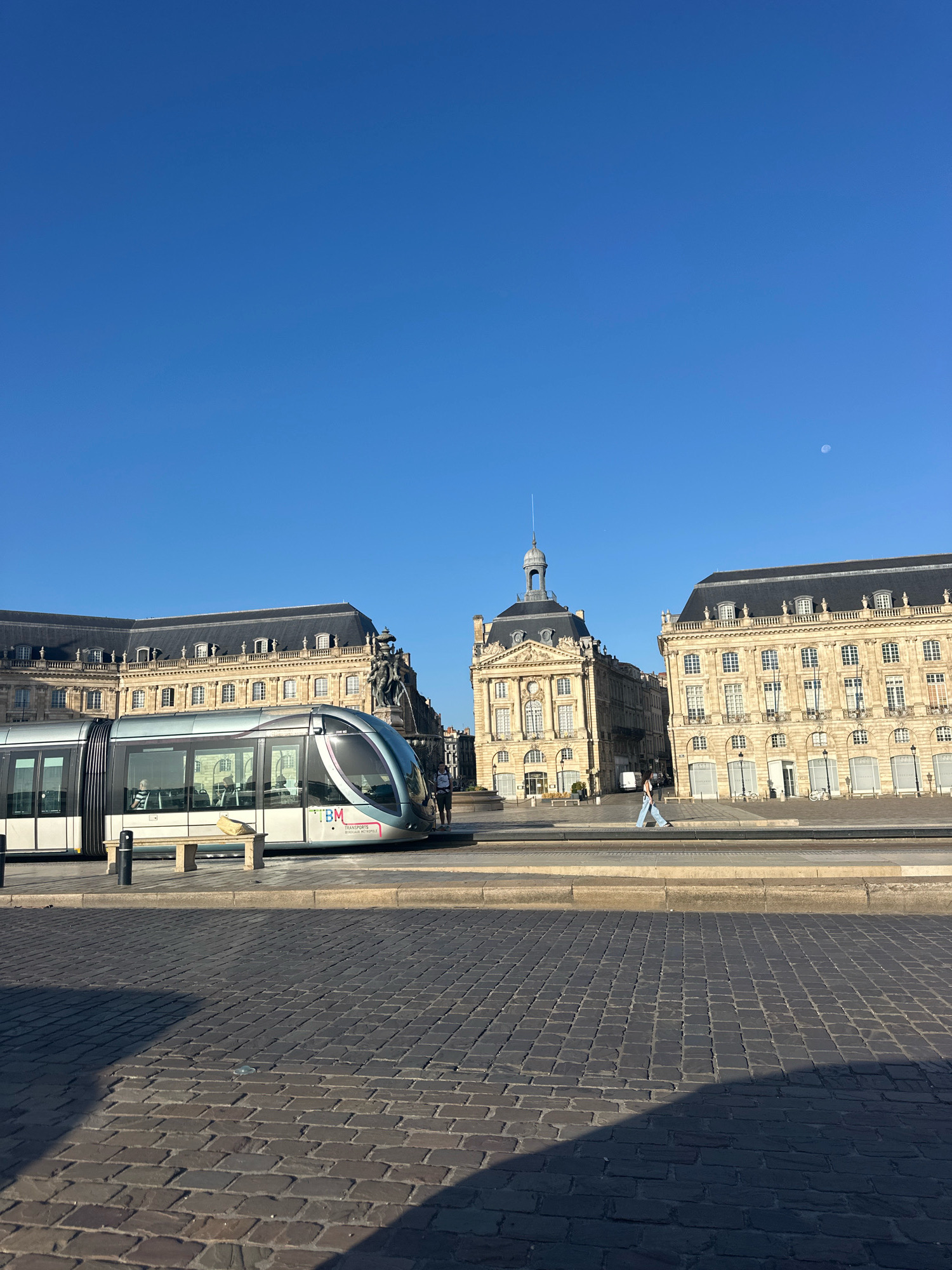 The image depicts a sunny day in Bordeaux, France, specifically at the Place de la Bourse. In the foreground, a cobblestone street is visible, with a shadow cast by an unseen object. To the left, a modern tram with a sleek, silver design is stopped. The tram has tinted windows, and a person can be seen sitting inside. The tram displays the branding "TBM TRANSPORTS BORDEAUX METROPOLE." Beside the tram, a concrete bench with a yellow bag on it is positioned on a raised platform.
In the background, the iconic Place de la Bourse, a grand 18th-century square, is prominently featured. The architecture is characterized by classical style buildings with stone facades, numerous windows, and dark mansard roofs. One building, the Customs House (Bureau des douanes), is particularly distinctive with its dome and central spire. A person is walking across the square in front of these buildings, dressed in light blue jeans and a light-colored top. The sky above is clear and a brilliant blue, with a small, pale moon visible in the upper right quadrant. The overall scene conveys a sense of urban tranquility and historical grandeur.