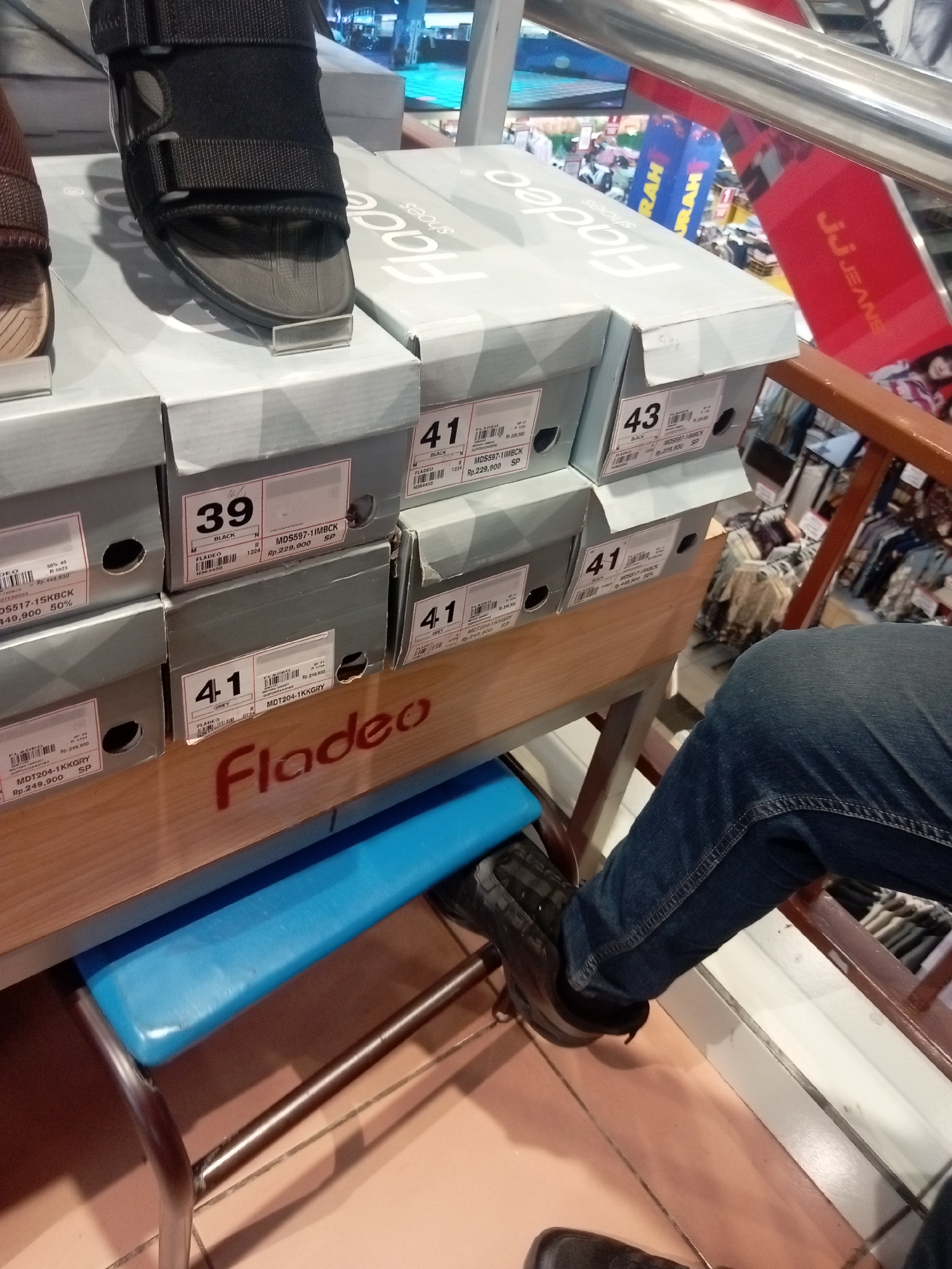 The image shows a retail display of footwear in a store located in Batam, Indonesia. The display features several shoe boxes stacked on shelves. The shoe boxes are predominantly grey with some branding visible, including the word "Fladeo". Some boxes have labels with shoe sizes and prices. One shoe box clearly displays the size "39" with the item code "MD5597-1MBCK". Another box shows size "41" with item code "MDT204-1KKGRY". A shoe from the brand "Fladeo" is placed on top of one of the boxes. In the foreground, a person's legs and feet are visible. The person is wearing dark blue denim jeans and black footwear. They are positioned in front of the display, suggesting they are a customer or an employee. The background reveals more of the store environment, including clothing racks and displays, indicating it's a large retail establishment. The overall scene depicts a typical shoe section within a department store or shoe shop in a commercial area of Batam.